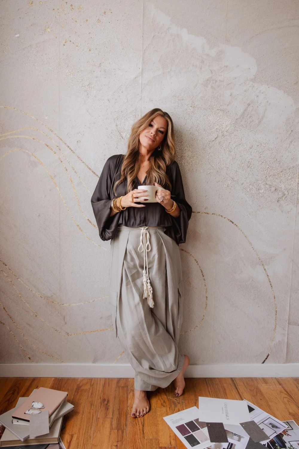 A woman with long wavy hair leaning against a wall, holding a white mug, wearing a dark satin blouse and loose beige pants, barefoot, with magazines and jewelry on the wooden floor.