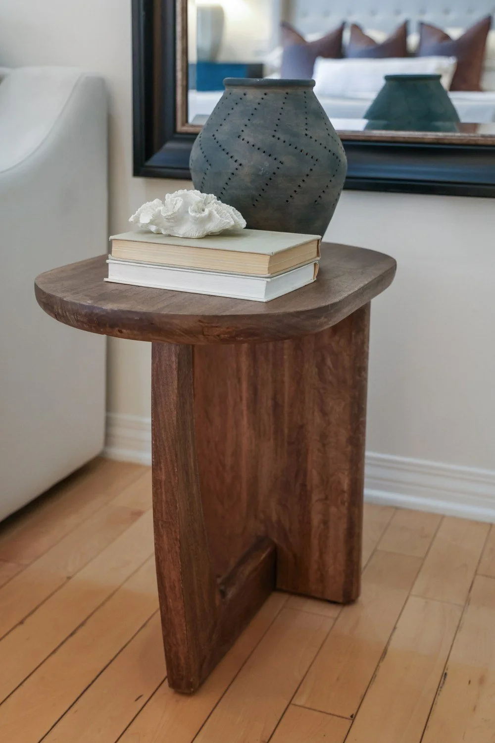 Decorative wooden side table with a ceramic vase, two books, and a piece of coral in front of a mirror.