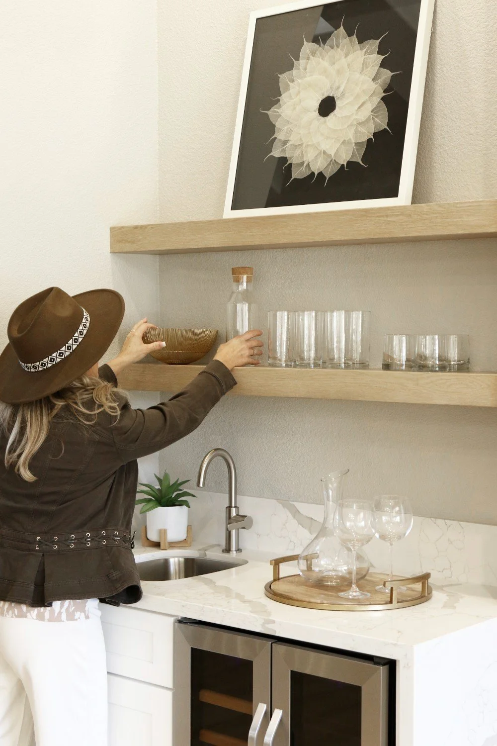 A woman with blonde hair wearing a brown hat and jacket arranges glassware on kitchen shelves above a white marble countertop.