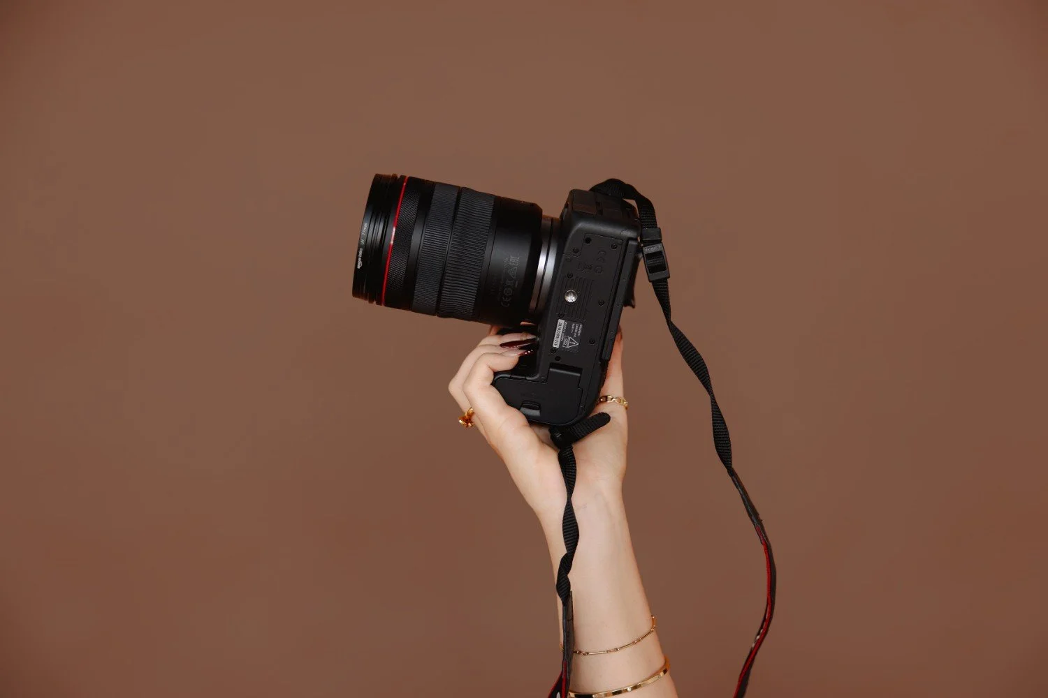 A person's hand holding a professional camera with a large lens against a plain brown background.