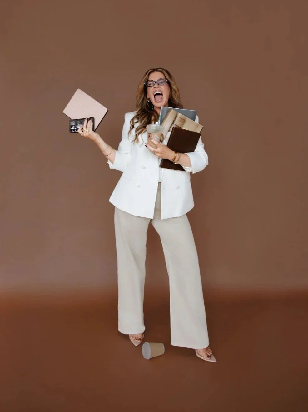 Woman in white business suit holding various books and drink, with a distressed expression, standing on brown background with a spilled coffee cup at her feet.