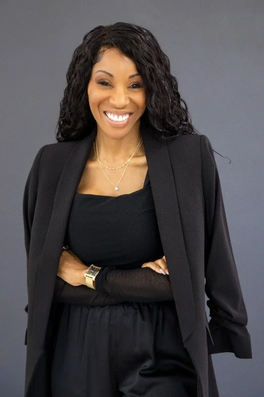 A woman with curly black hair smiling, wearing a black dress and blazer, gold jewelry, and a gold watch, standing against a gray background.