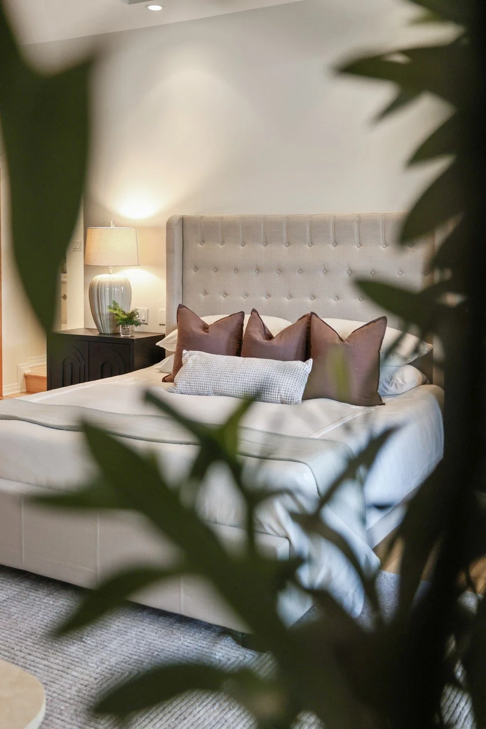 A neatly made bed with a tall, upholstered headboard, accented by multiple brown pillows and a smaller white pillow, in a cozy bedroom with a nightstand holding a table lamp and a small plant, viewed through green leaves.
