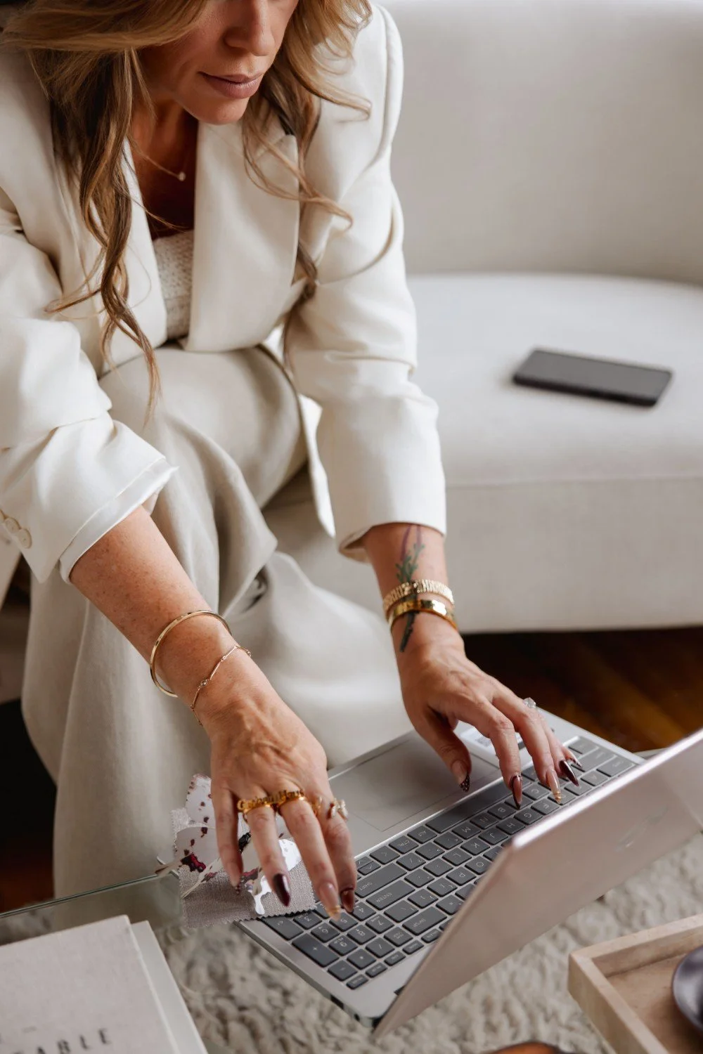 Woman in white blazer using a laptop on a glass table, with jewelry and tattoos on her hands.