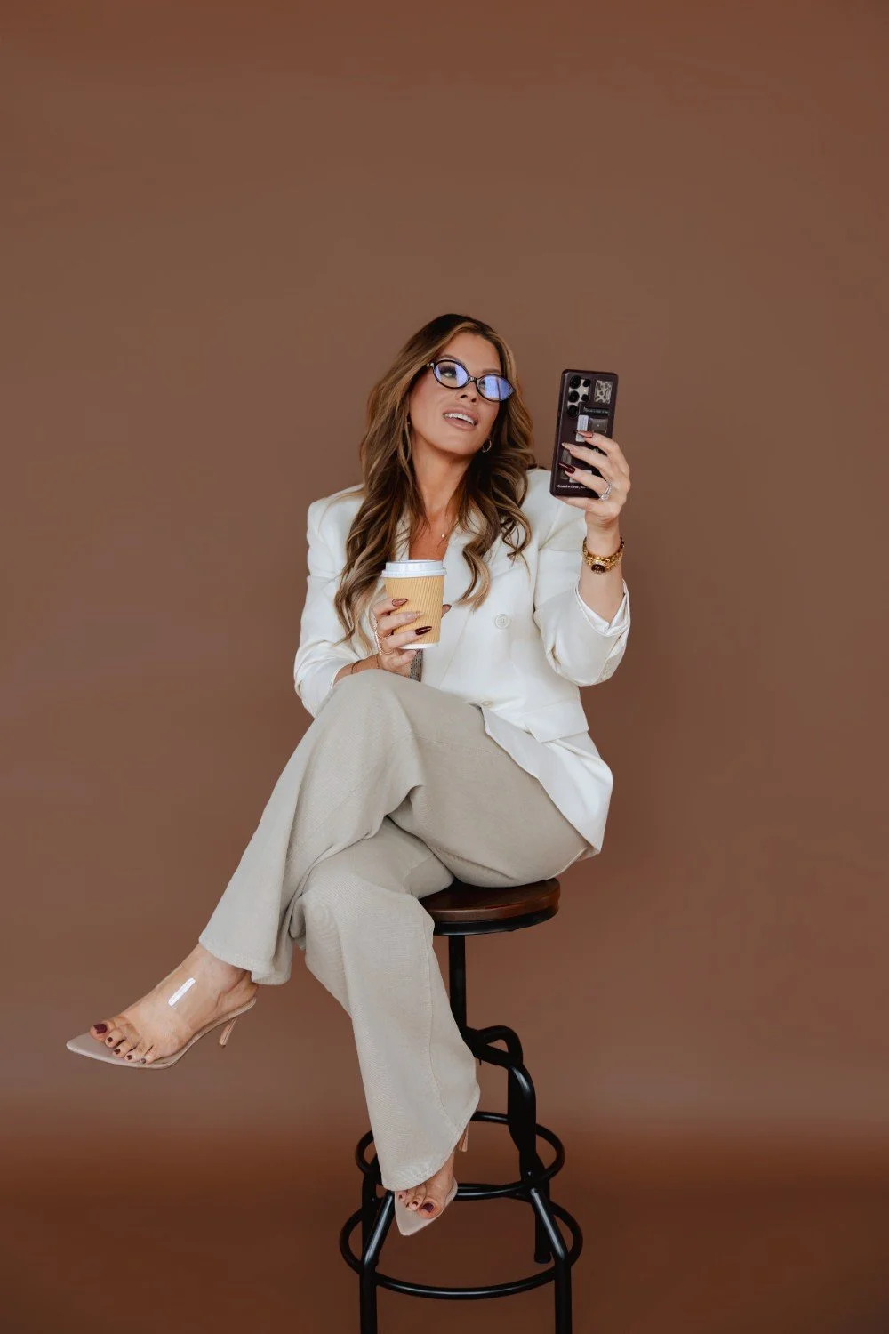 A woman with wavy brown hair wearing glasses, a white blazer, beige pants, and nude high heels is sitting on a black stool against a brown background. She is holding a to-go coffee cup in one hand and taking a selfie with her phone in the other.