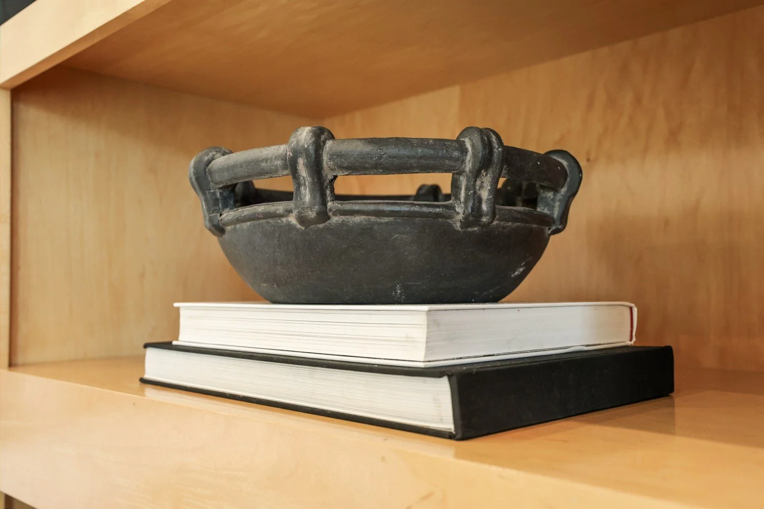 A black bowl with a decorative handle sitting on top of two stacked books on a wooden shelf.