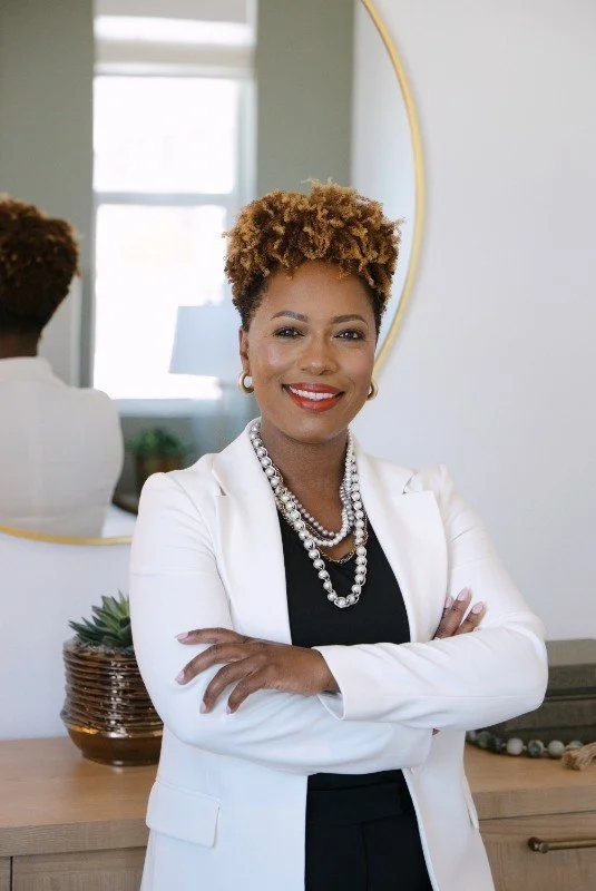 A confident woman with natural curly hair and dark skin, wearing a white blazer and pearl necklace, standing in a modern, well-lit room with a mirror and decorative plant in the background.