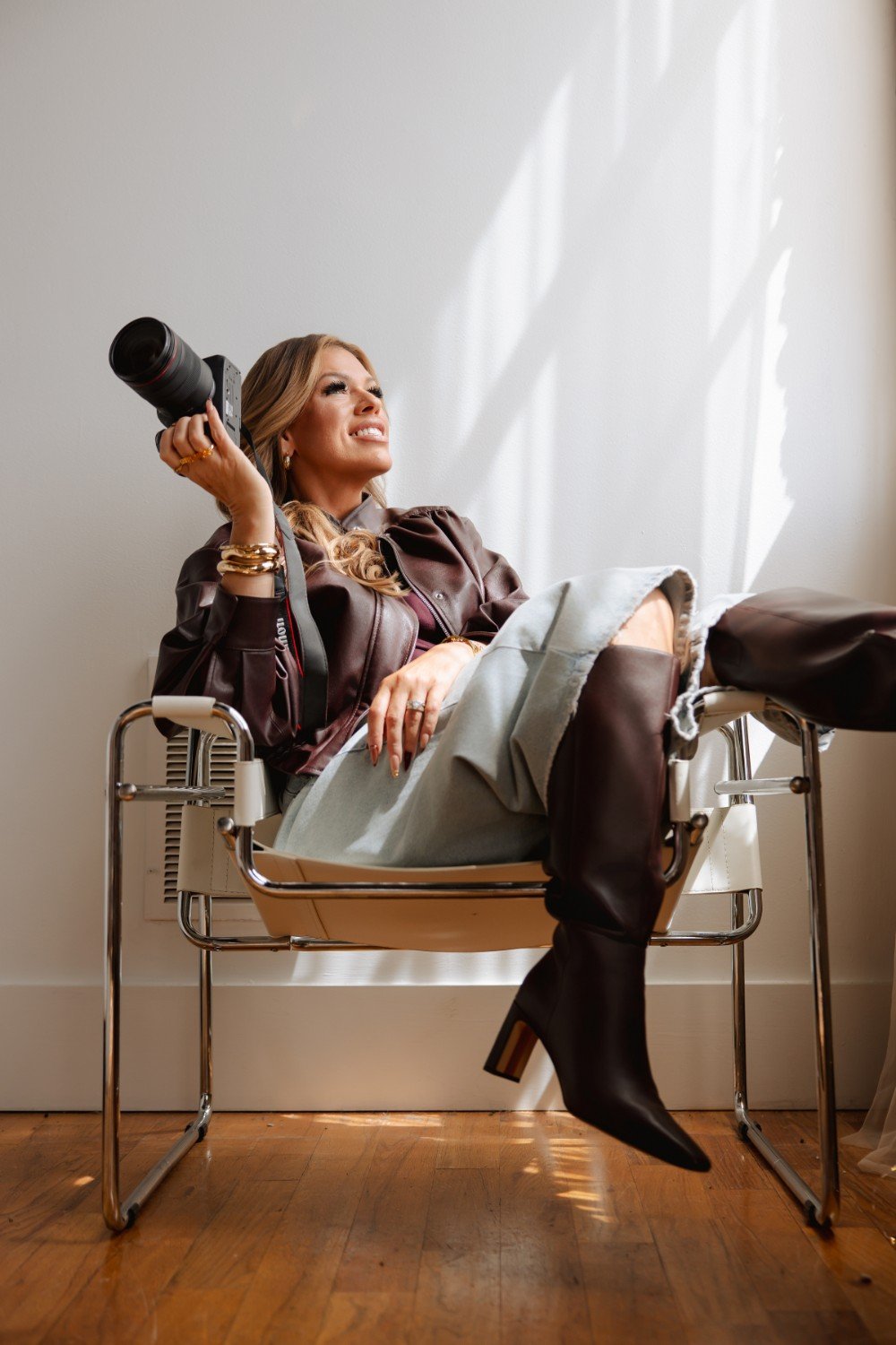 A woman sitting on a modern metal chair, smiling and holding a professional camera. She wears a brown leather jacket, a skirt, black high-heeled boots, and gold jewelry. Light from a window creates shadow patterns on the wall.