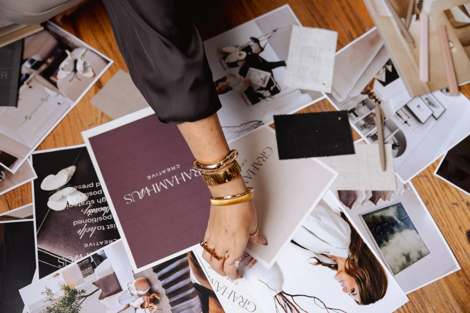 A person's hand with gold jewelry examining interior design mood boards, fabric samples, and photographs spread out on the floor.