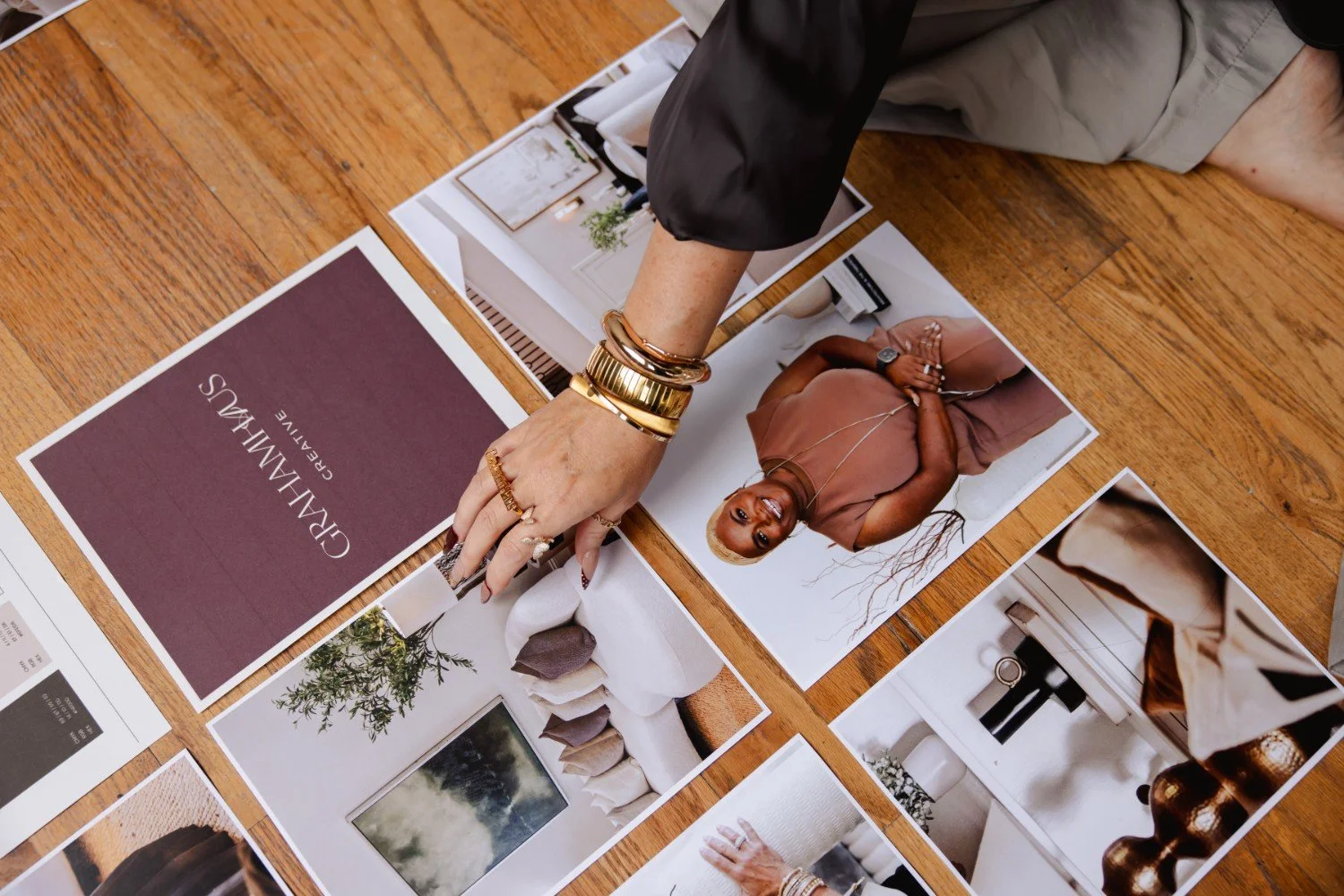 A person with multiple gold and jewelry bracelets is arranging interior design photos and a branding card on a wooden floor, with furniture and decor images.