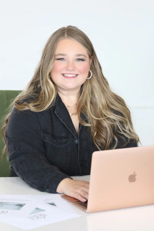 A young woman with long blonde hair, wearing a black denim jacket and gold hoop earrings, smiling while sitting at a white desk with a pink laptop and papers.