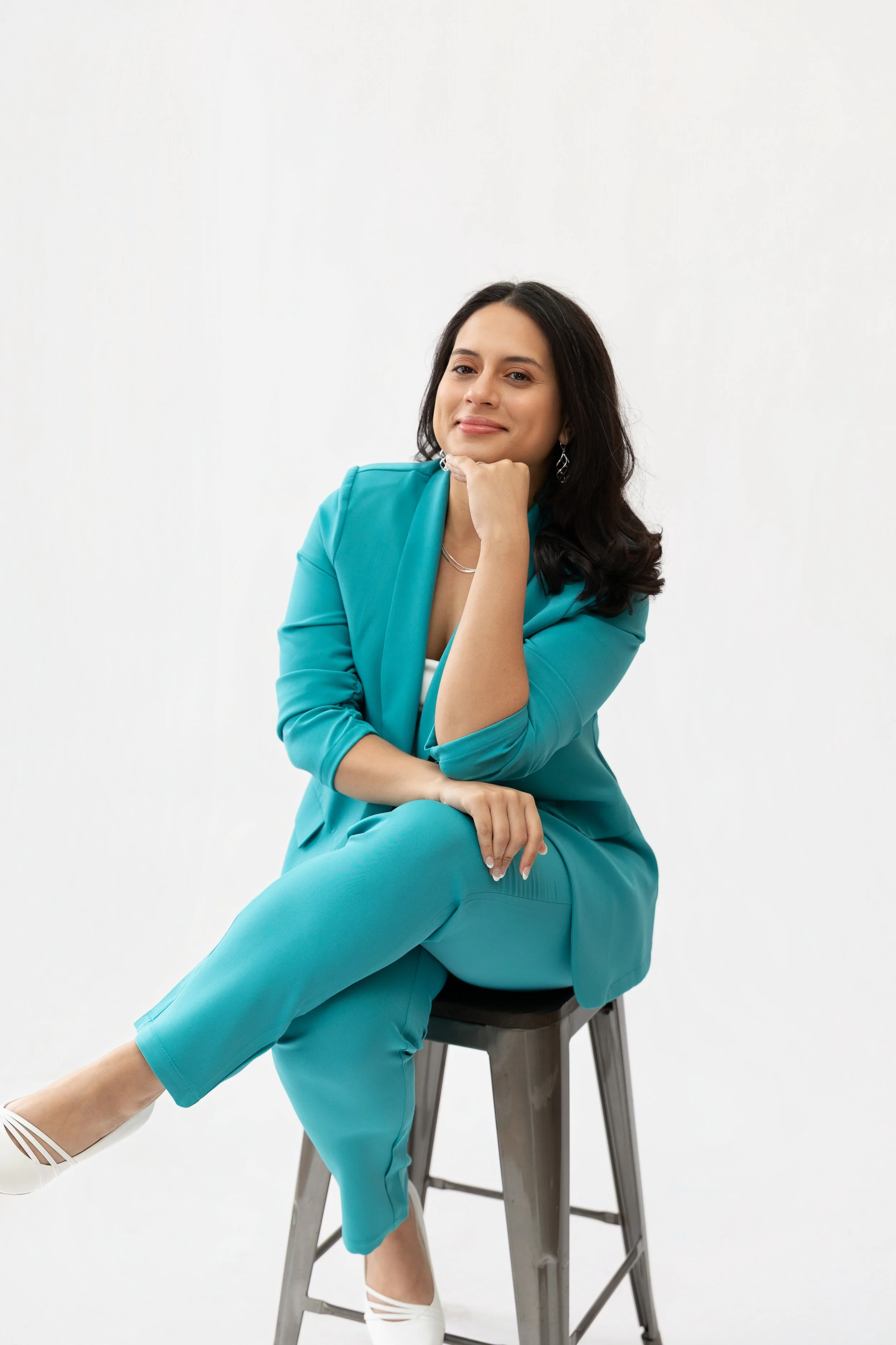 A woman with dark hair styled in loose waves, wearing a teal blazer and matching pants, sitting on a stool against a plain white background. She has a smile on her face, her left hand resting on her chin, and her right hand on her knee. She’s dressed with earrings and a necklace, and wearing white high heels.