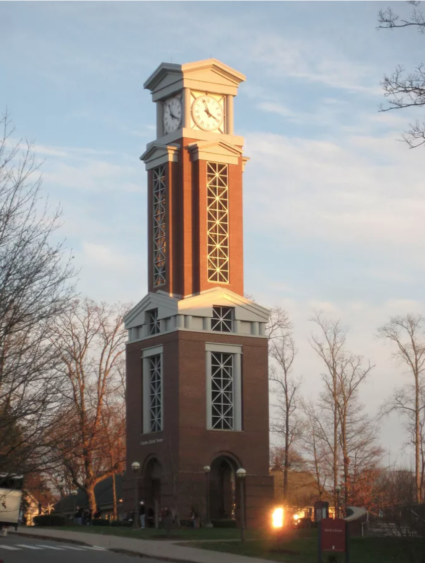 A tall brick clock tower with white decorative elements, displaying the time at around 4:15, surrounded by leafless trees and a few people at the base during sunset.