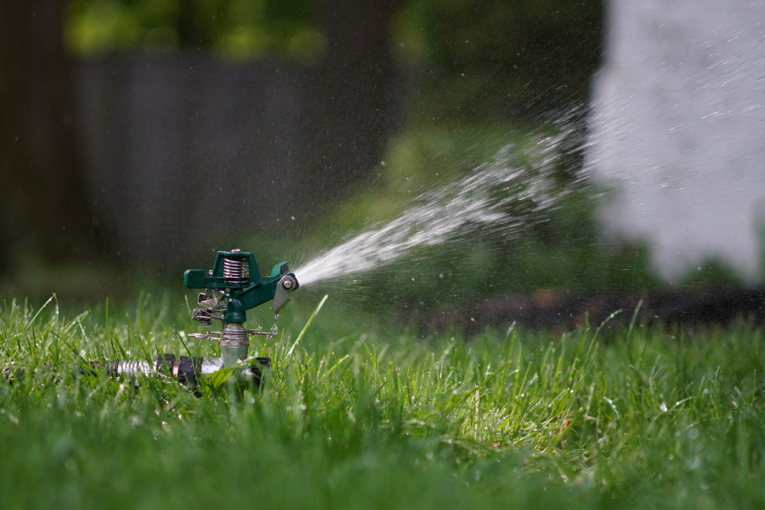 A close-up image of a green garden sprinkler spraying water across a grassy lawn with blurred trees and bushes in the background.