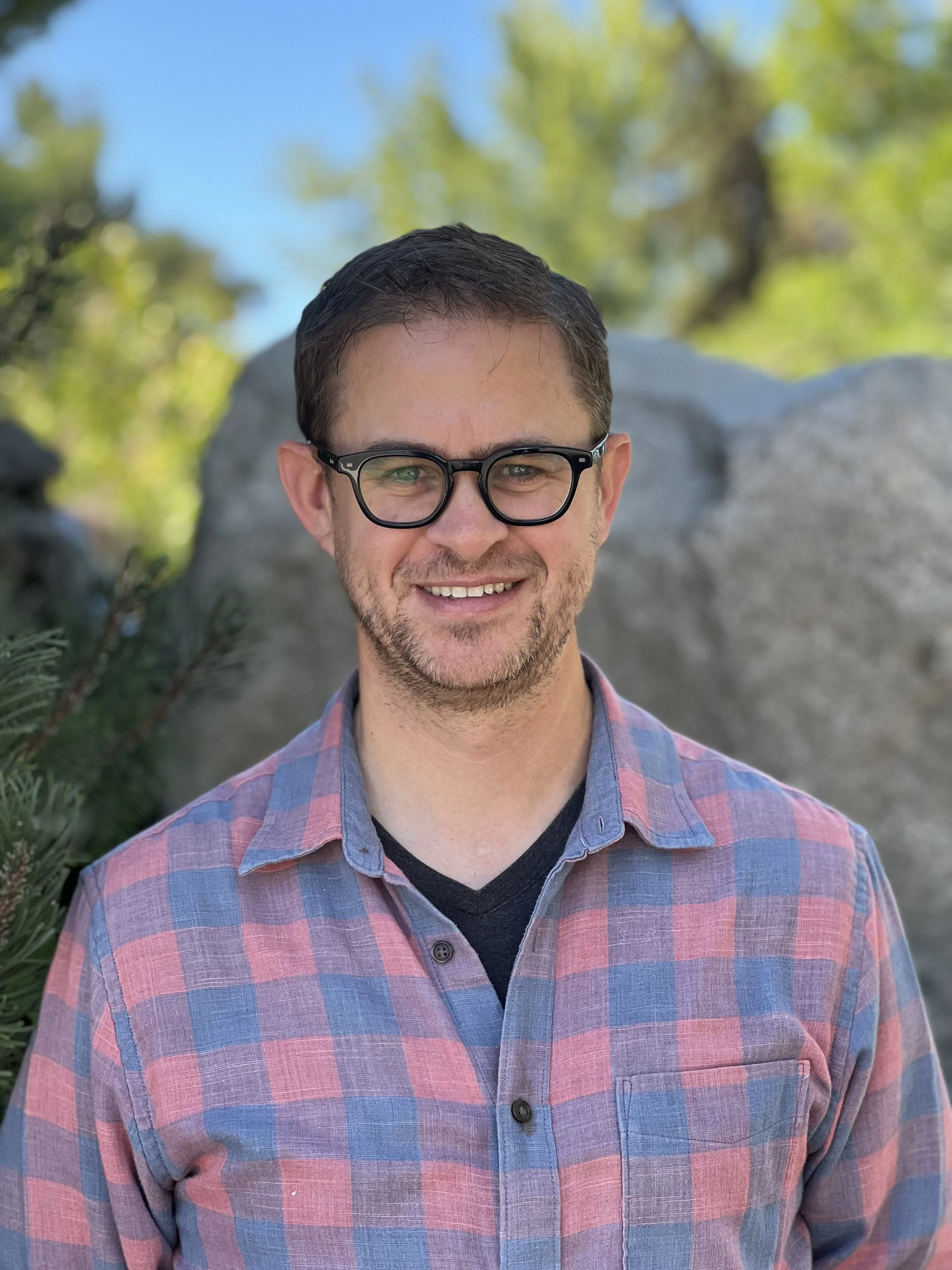 A man wearing glasses and a plaid shirt with a dark t-shirt underneath, smiling outdoors with trees and rocks in the background on a sunny day.