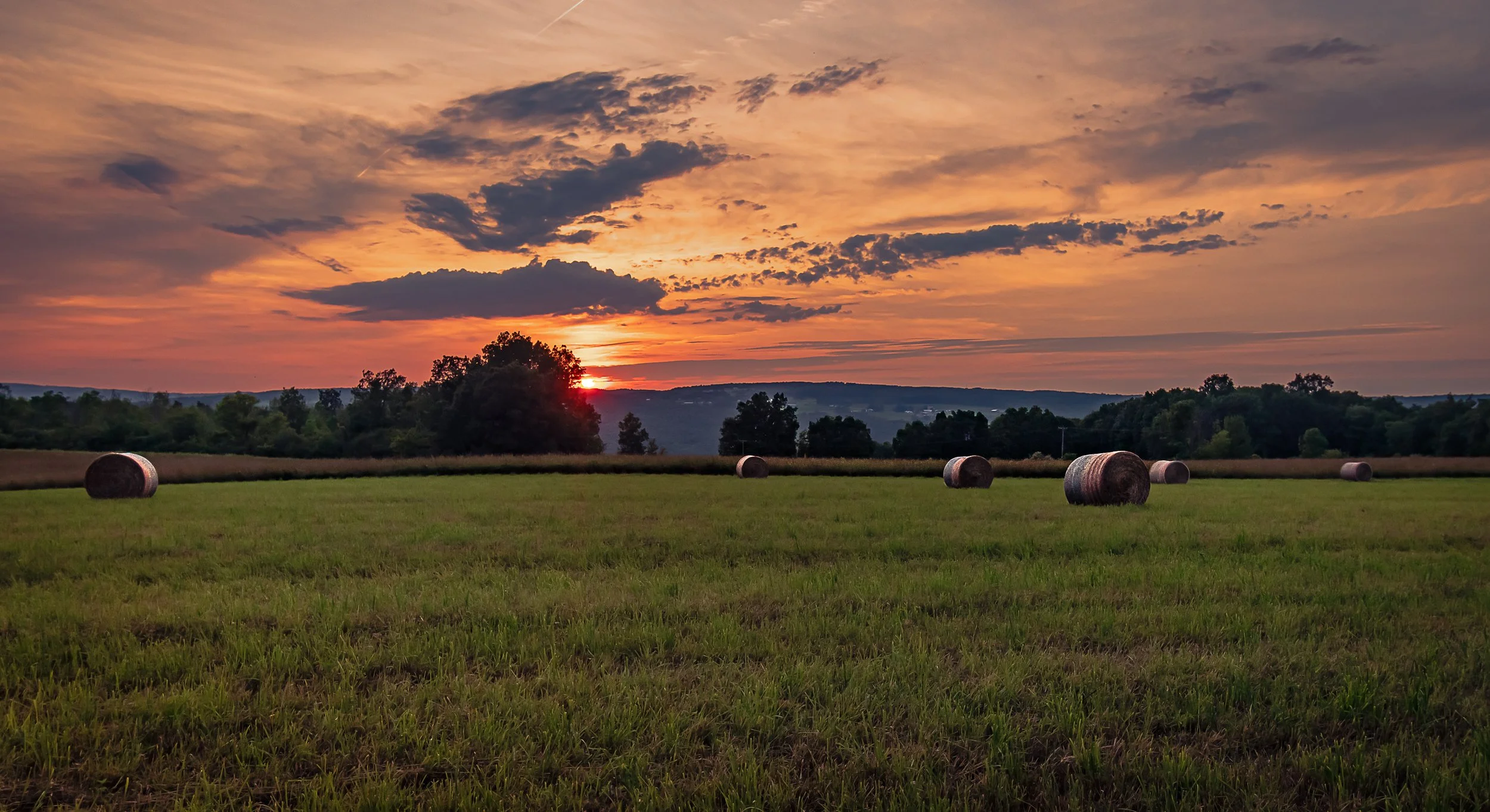A pastoral scene at sunset with a colorful sky, trees on the horizon, and a grassy field with several hay bales scattered across the landscape.