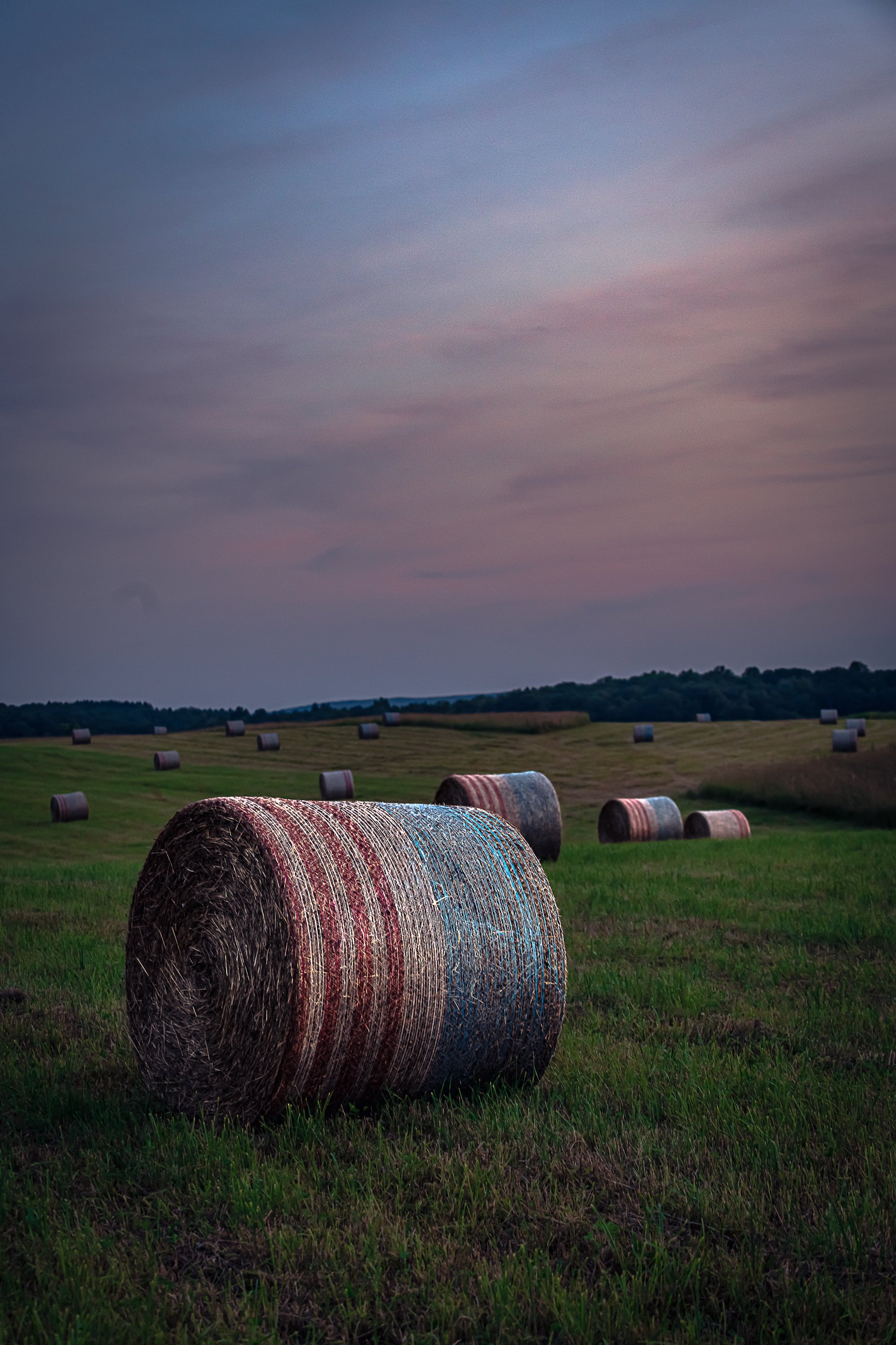 A field with large hay bales scattered across the green grass at dusk, with a colorful sky and hills in the background.
