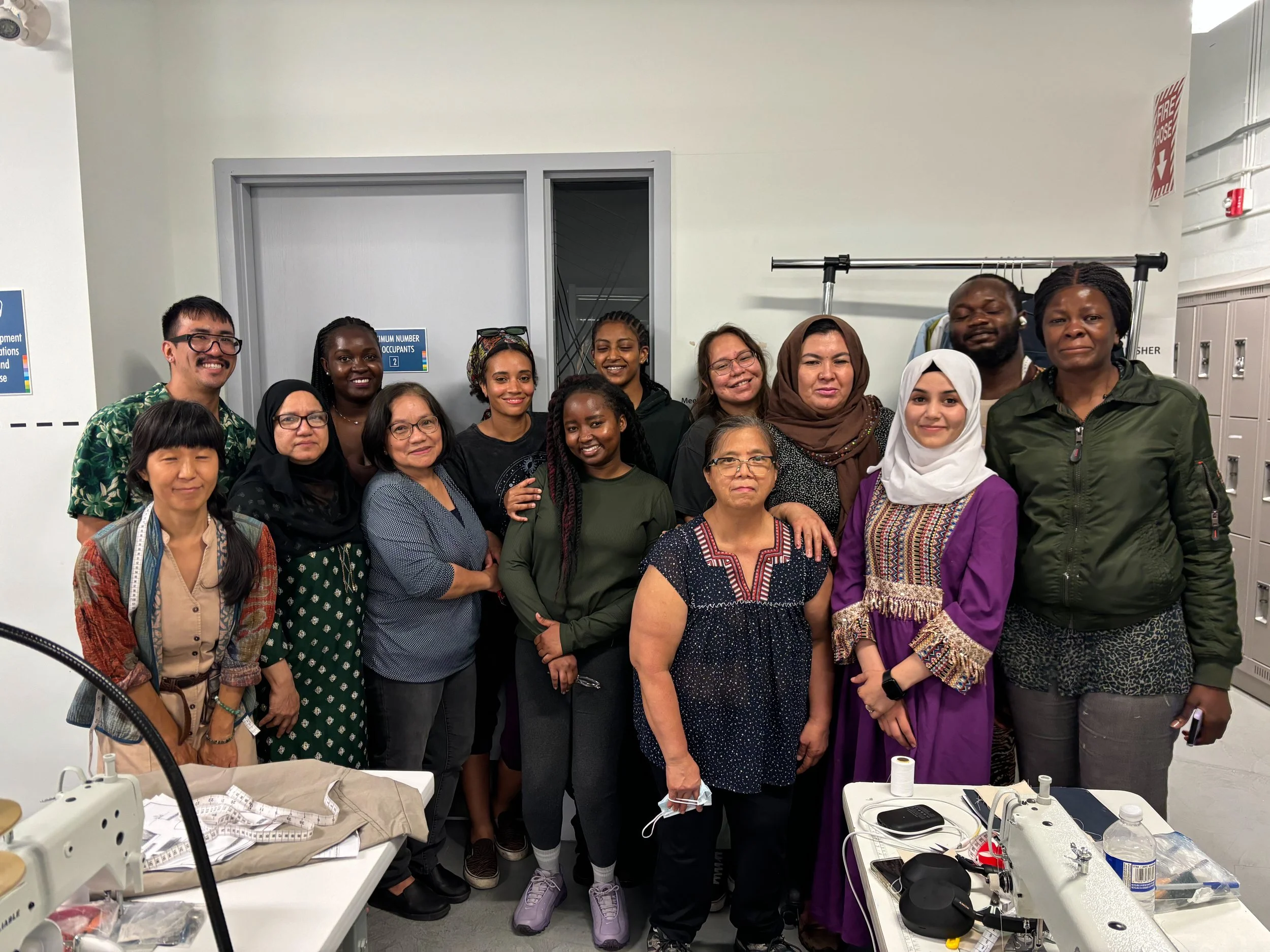 Carlos with his Industrial Power Sewing class. The classroom has sewing and tailoring supplies and equipment on tables, including measuring tape, fabric, and sewing tools.