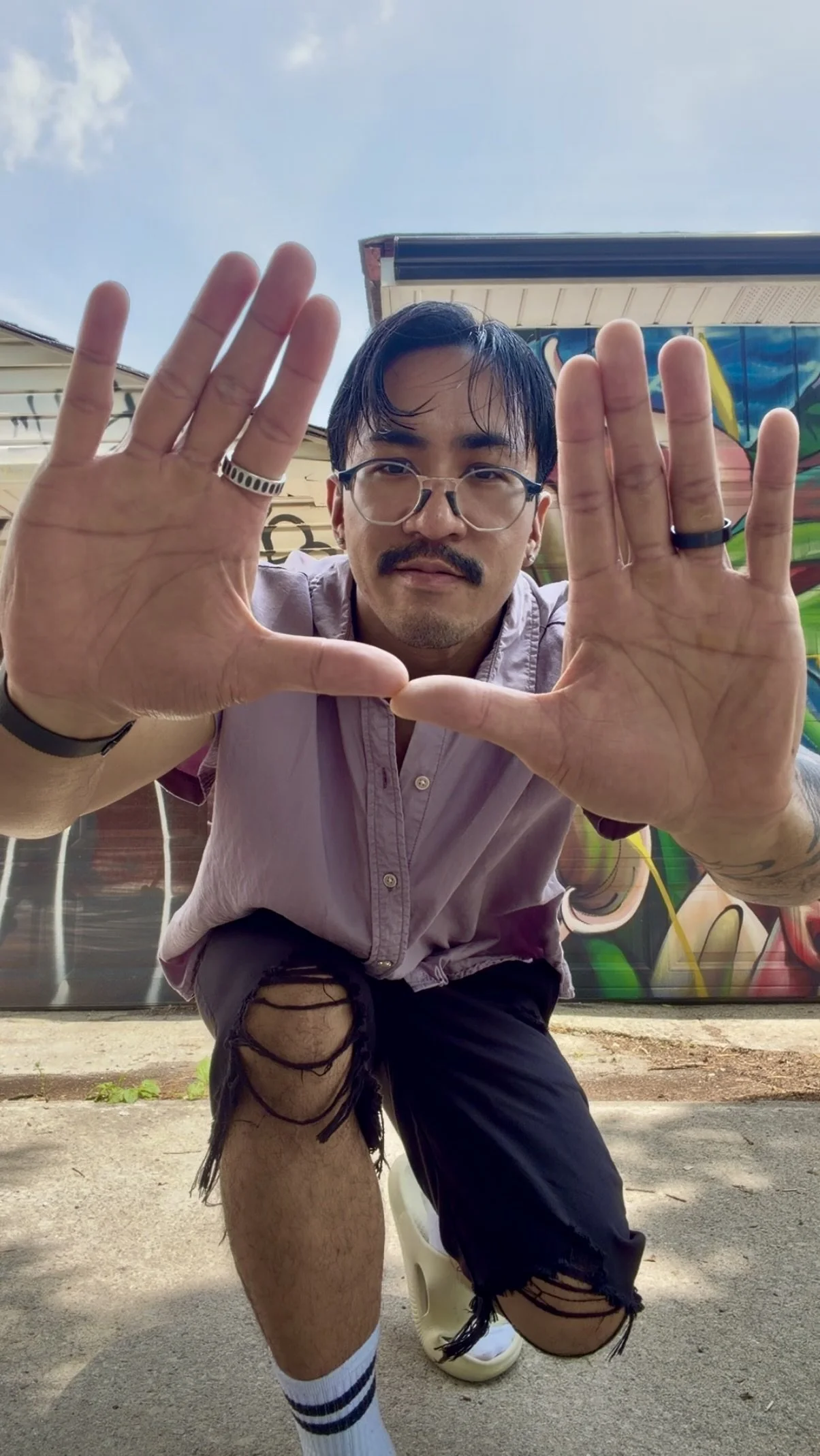 Carlos in Trinity Bellwoods Park, wearing a light purple shirt, ripped black shorts, and striped socks, in front of a colourful graffiti wall, framing the photo with his hands.