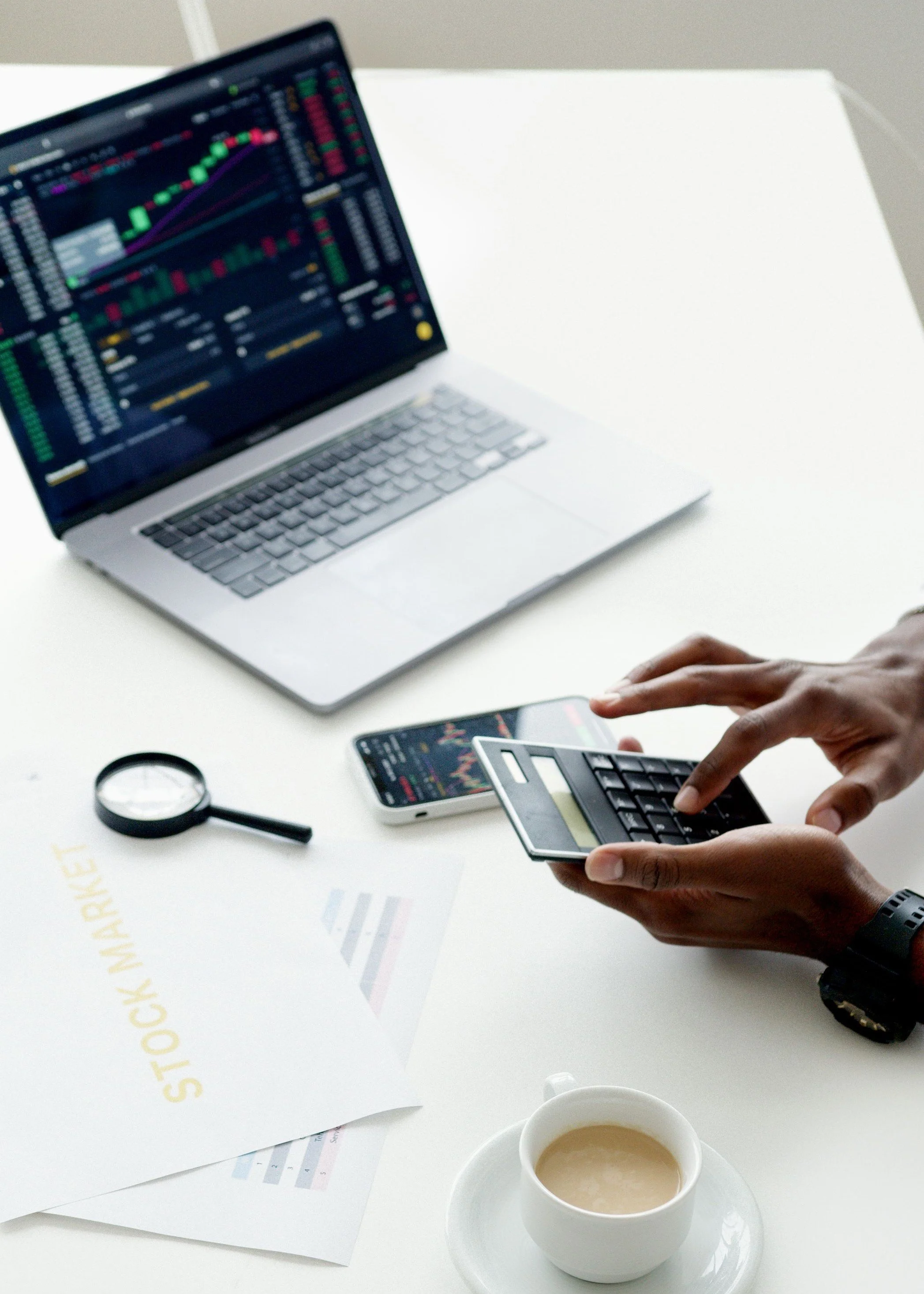 Una computadora portátil con gráficos y datos en la pantalla, una mano usando una calculadora, un teléfono móvil con gráficos, una lupa, y una taza de café en un escritorio blanco.