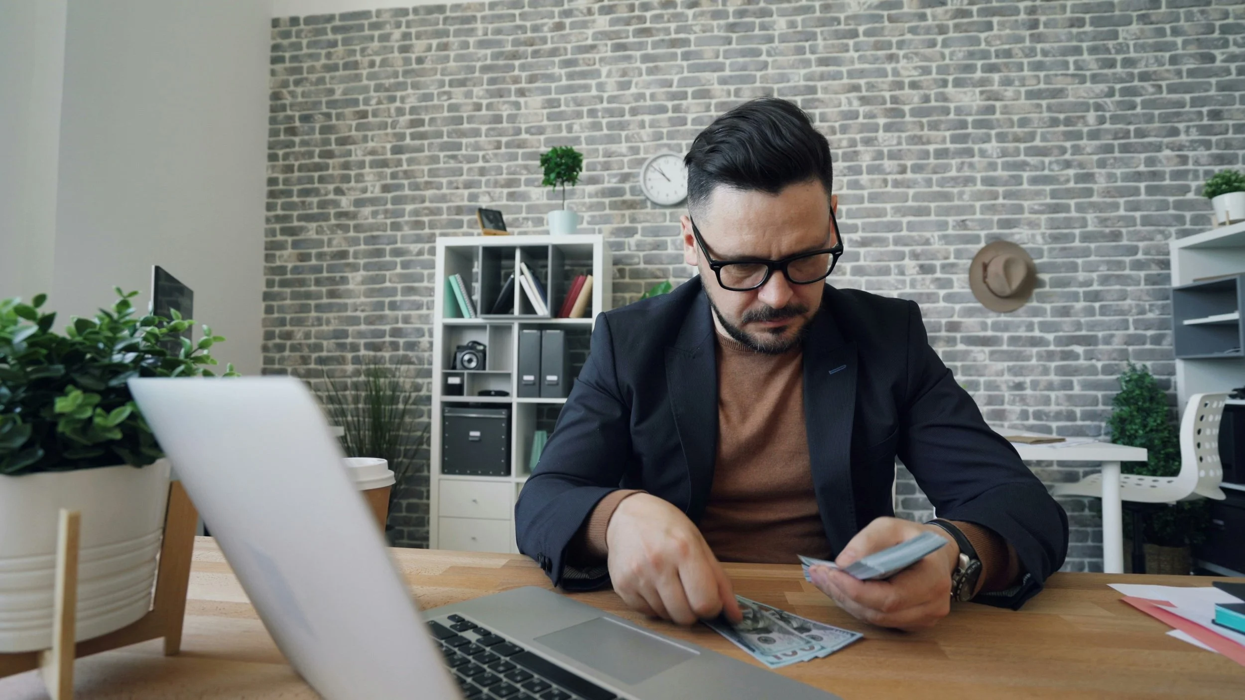 Hombre con gafas sentado en una oficina moderna, revisando dinero y usando su teléfono móvil, con estantería de libros y plantas en el fondo.