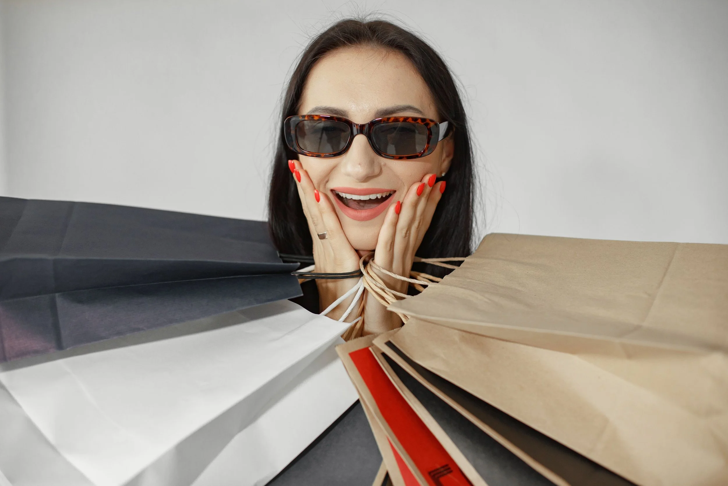 Mujer con gafas de sol grandes, sonriendo con manos en las mejillas, rodeada de bolsas de compras.