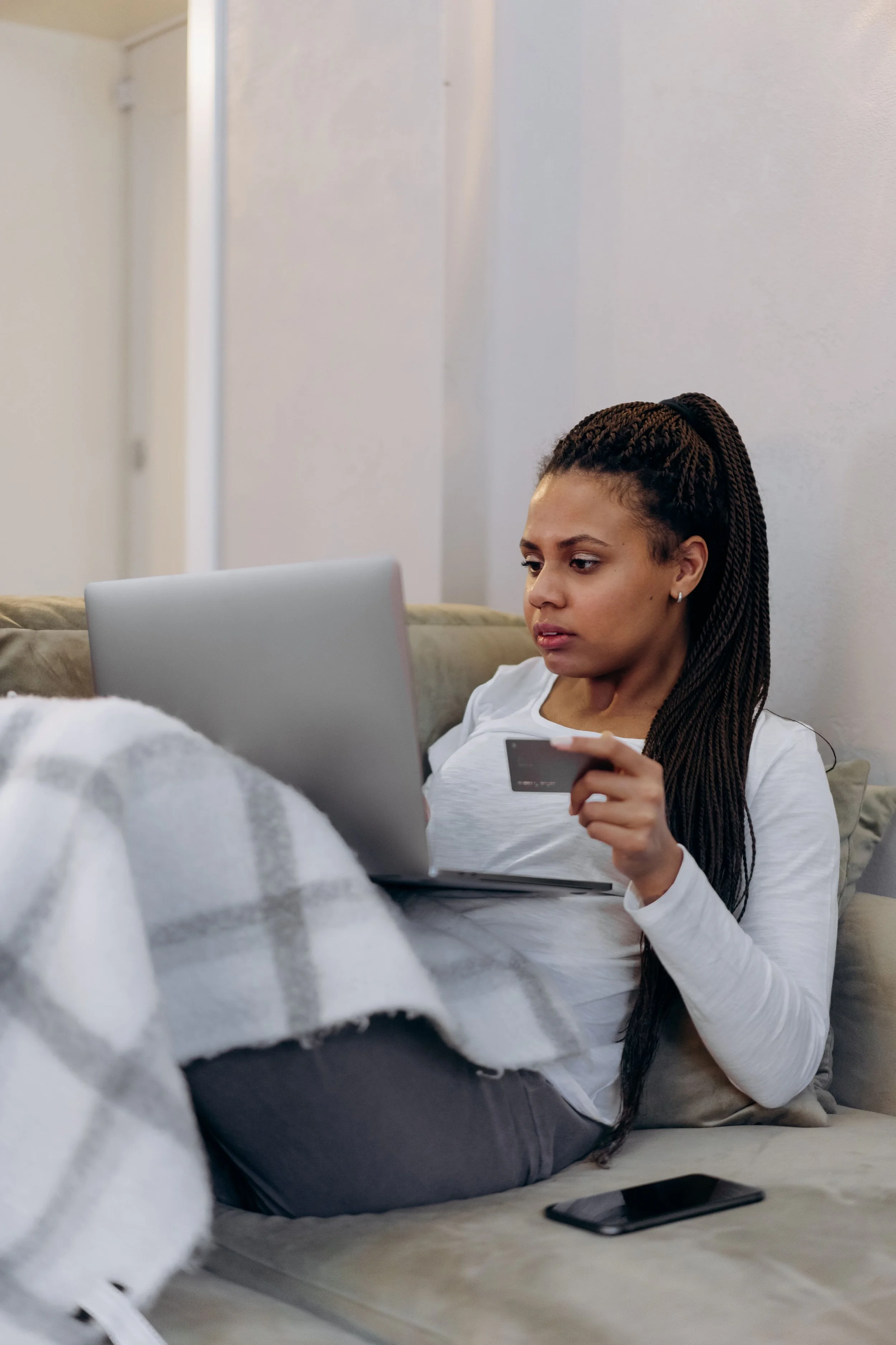 Joven mujer con trenza larga usando camiseta blanca, descansando en el sofá, con laptop y teléfono, en ambiente interior.