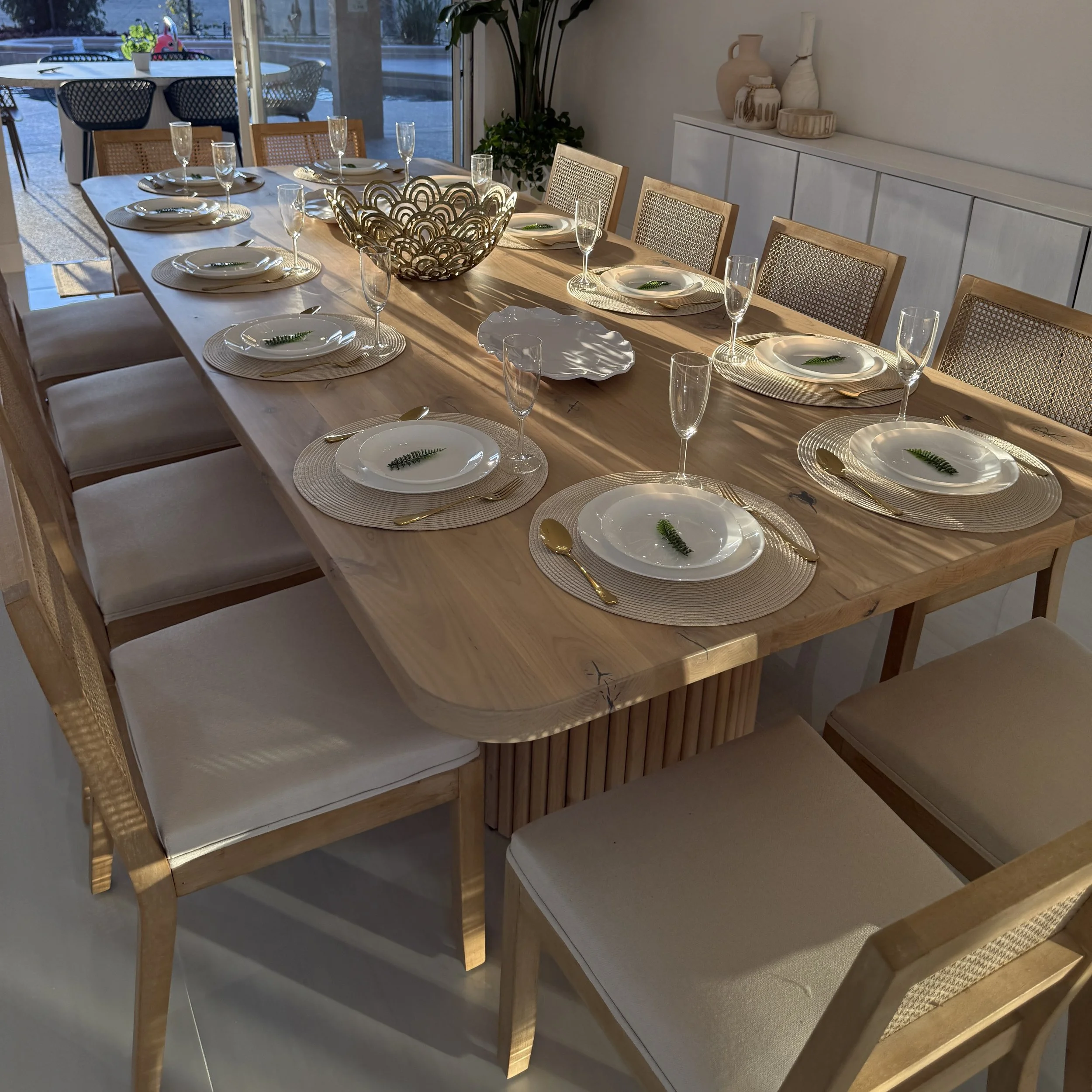 A dining table set for eight with cream-colored plates, gold utensils, and champagne flutes. The table has a decorative metallic bowl in the center. There are wooden chairs with woven backs and cushioned seats. A sideboard with white pottery and a large potted plant is in the background. Large windows show an outdoor patio with additional seating and a view of trees and a parking lot.