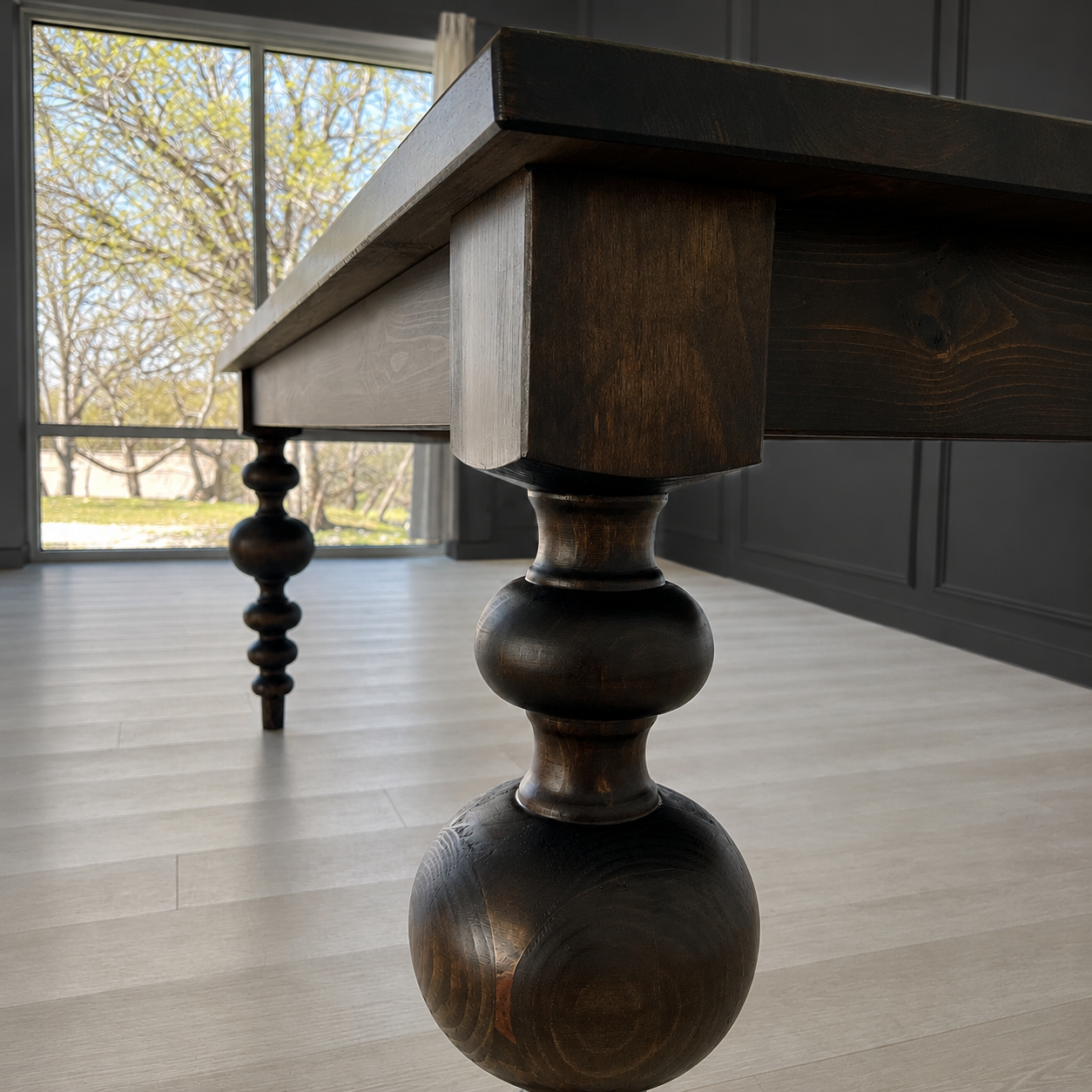Close-up of a dark wooden table with turned legs near a large window with trees outside and light-colored flooring.