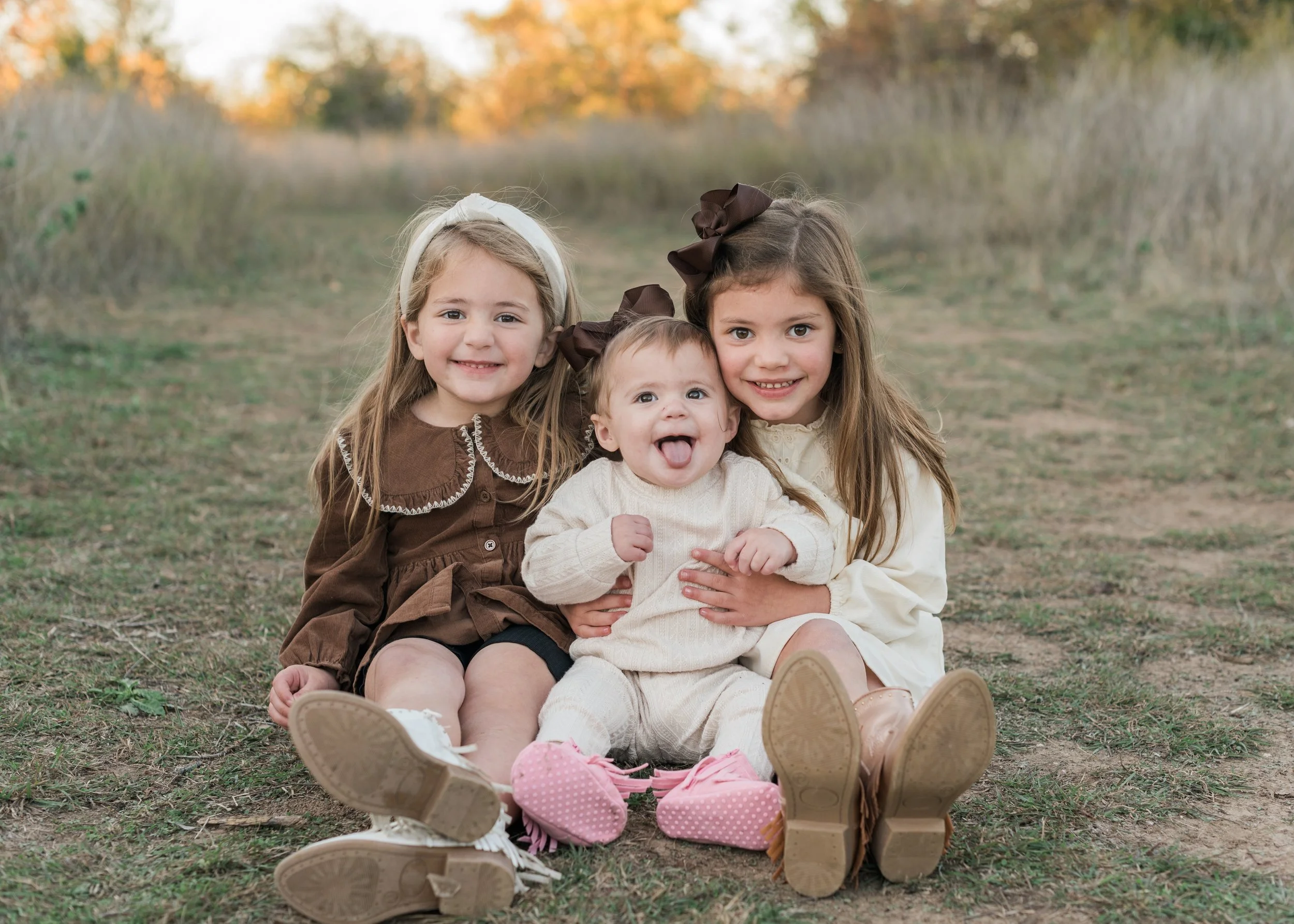 Four young girls sitting outdoors on the grass during autumn, smiling and looking at the camera. They are dressed in warm clothing, with two girls wearing large bows in their hair, and one girl making a funny face with her tongue out.