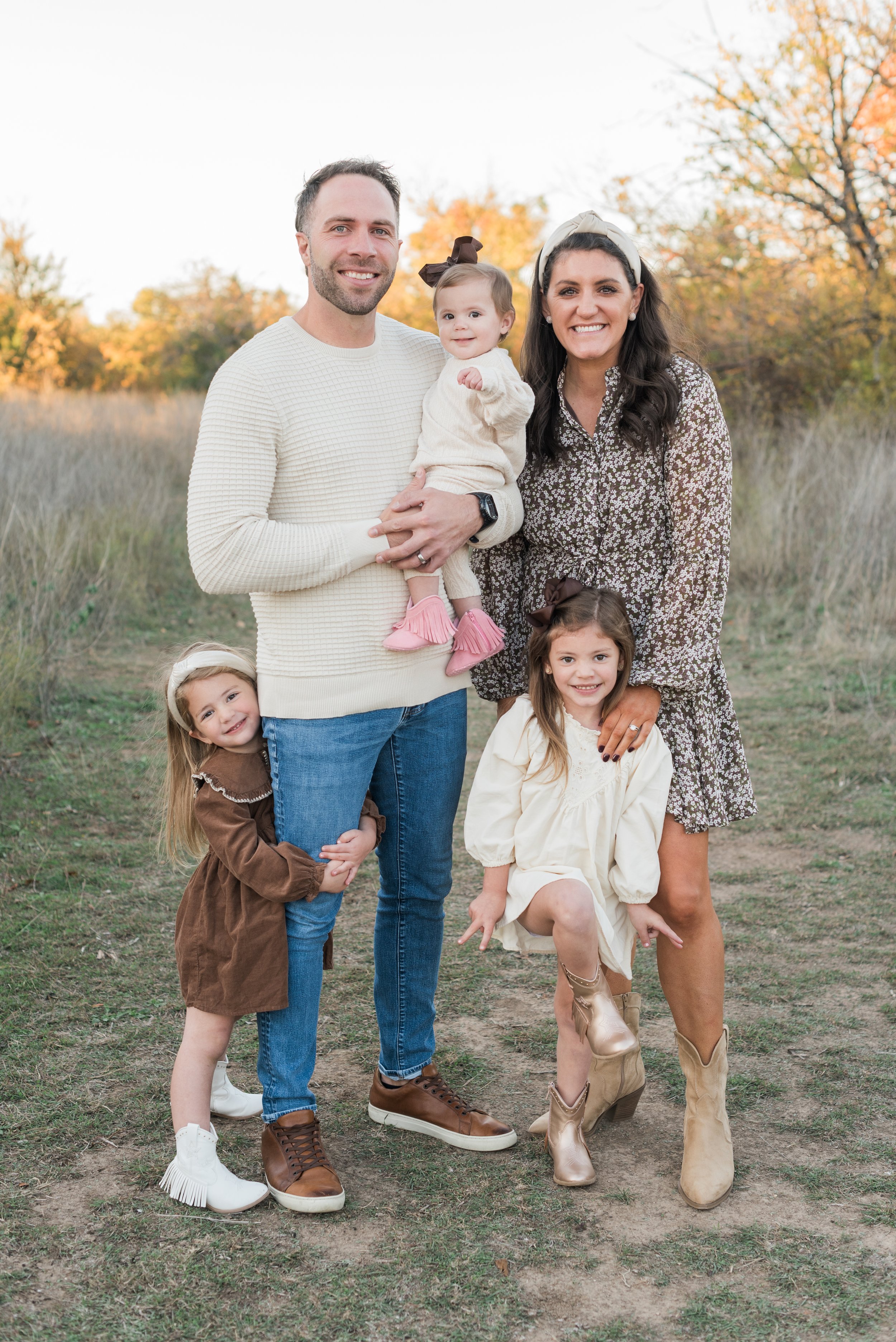 A family of six posing outdoors in a field during autumn. The family includes two adults, three young girls, and a toddler. The adults are smiling, and the children are in various playful poses.