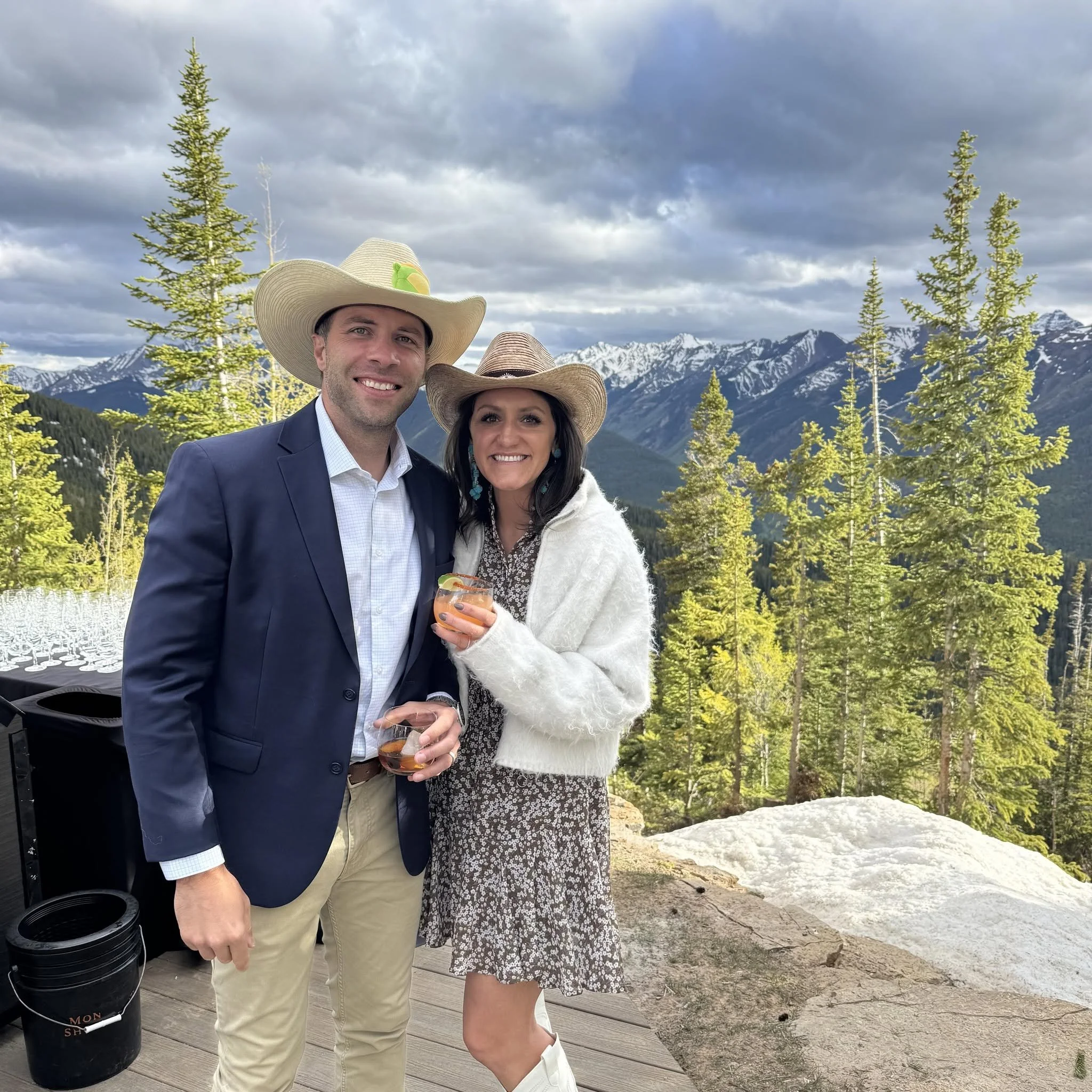 A man and woman smiling outdoors with mountains and trees in the background, both wearing wide-brimmed hats and holding drinks.
