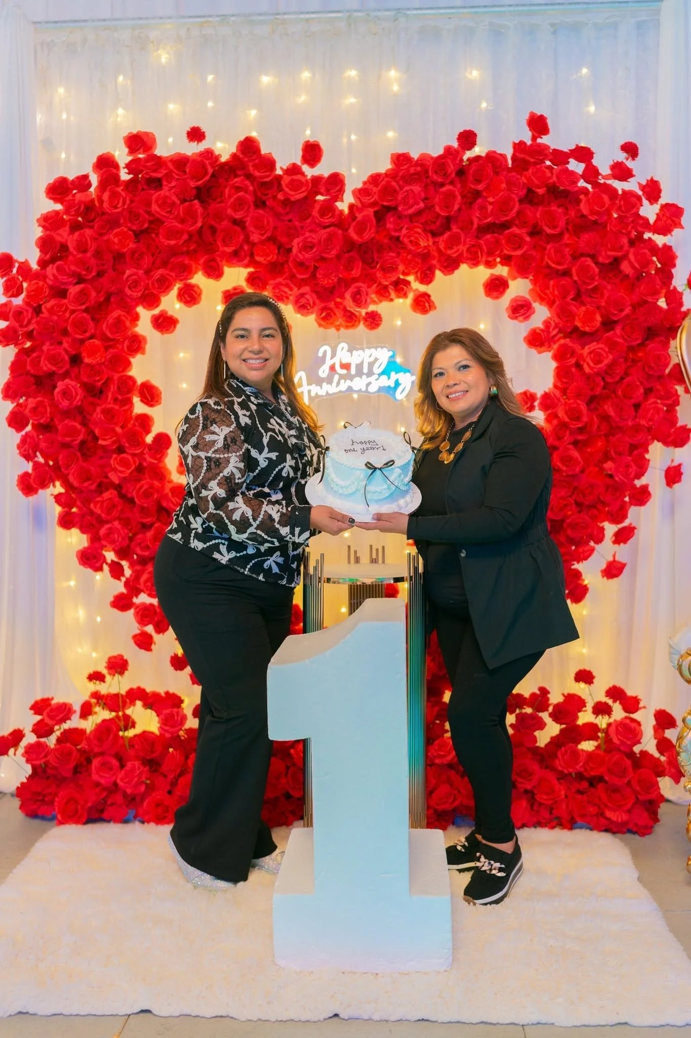 Two women are standing in front of a heart-shaped floral backdrop made of red roses, with a white curtain and string lights behind them. They are smiling and holding a cake with a message, celebrating an occasion.
