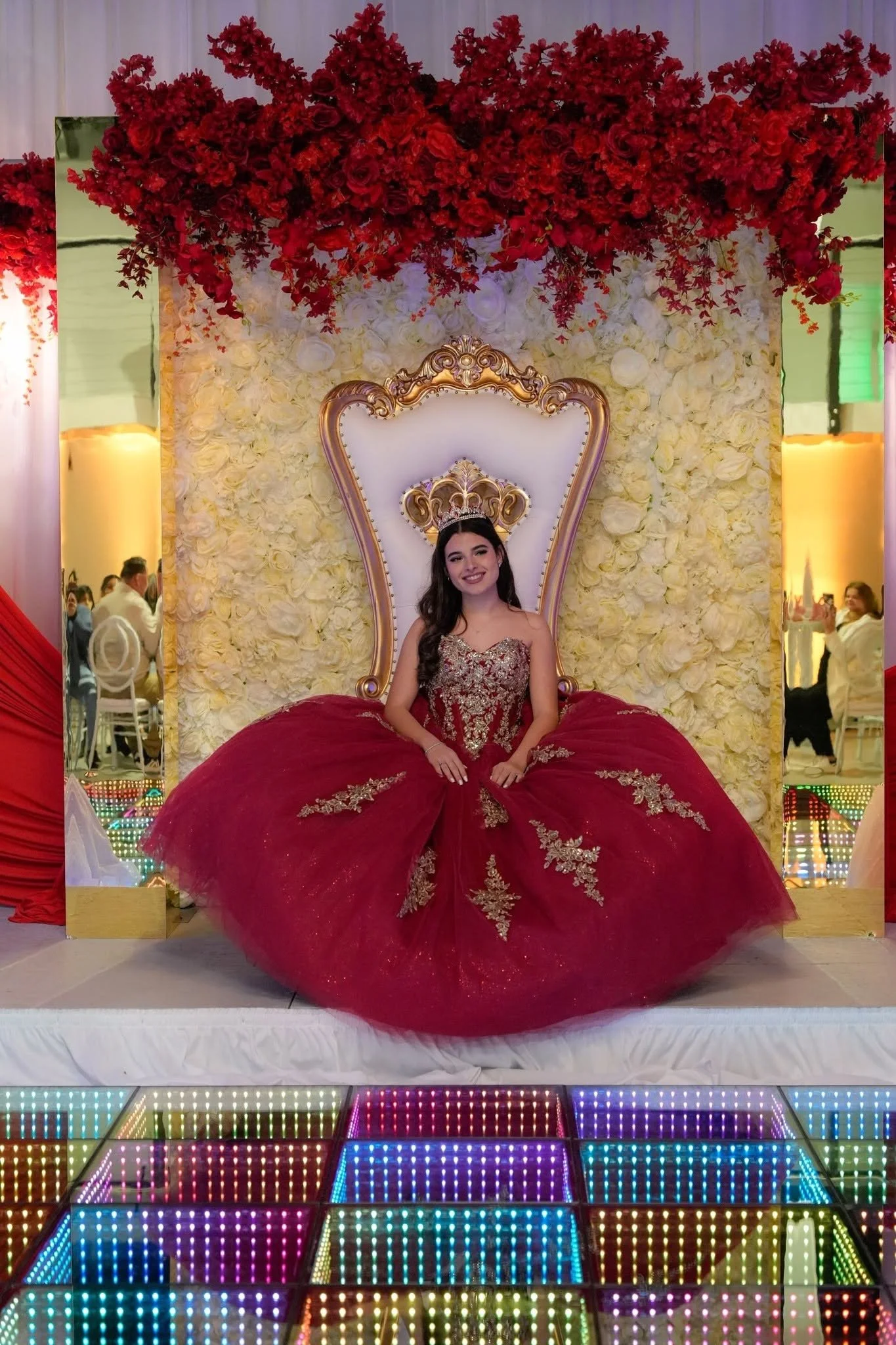 A young woman wearing a tiara and a glamorous red gown with gold embroidery is sitting on an ornate white and gold throne. The backdrop is decorated with white flowers and overhead red floral arrangements. The floor features colorful LED lights.