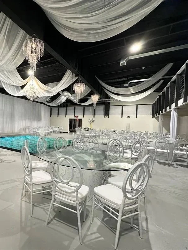 Elegant indoor event space decorated with white drapery and chandeliers, featuring a swimming pool and white round tables with white chairs.