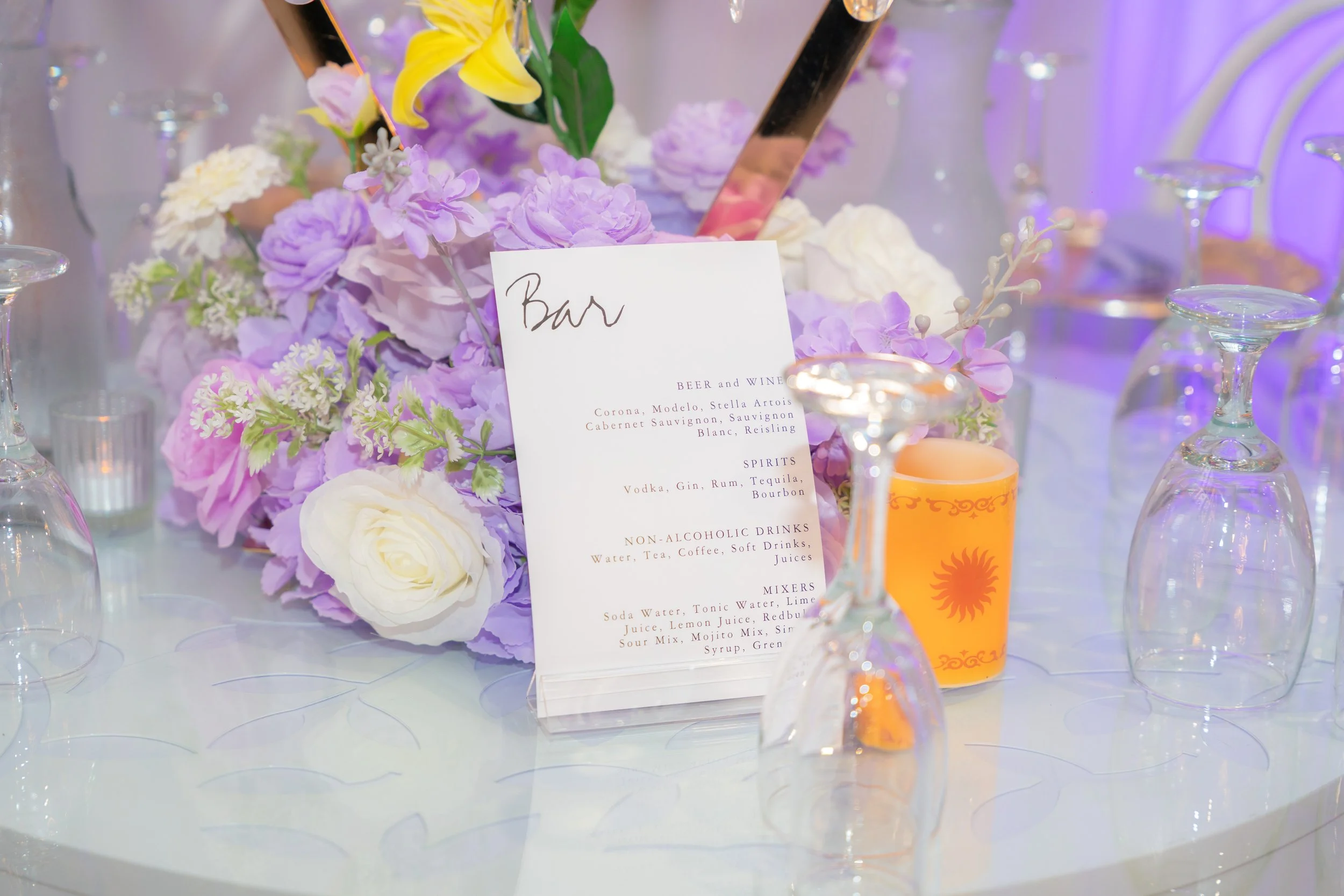 A floral centerpiece with purple, white, and yellow flowers on a table, surrounded by empty wine glasses and a drink menu. The background has soft purple lighting.