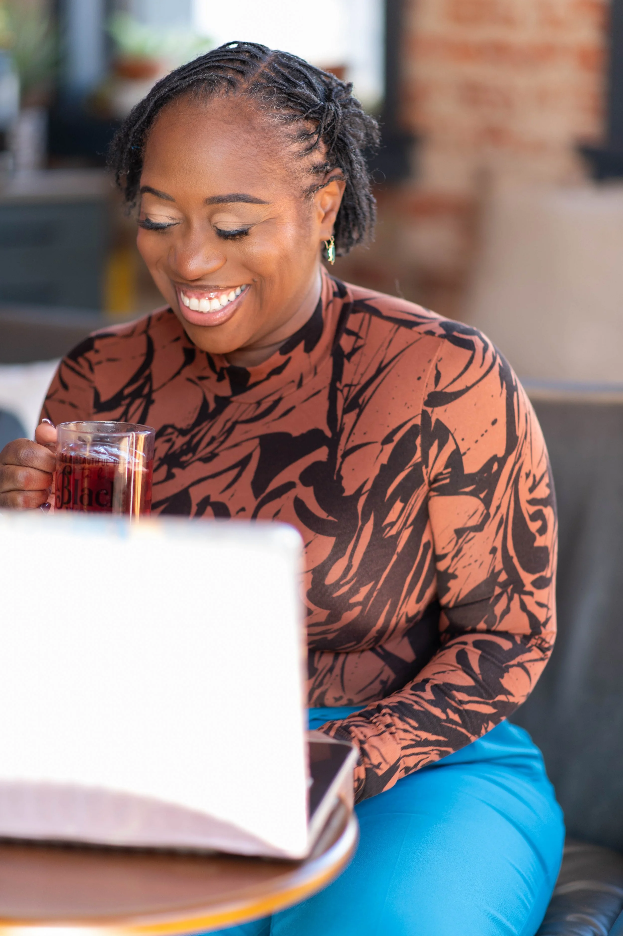 A smiling woman with braided hair and earrings, wearing a patterned brown and black top and blue pants, sitting at a table with a glass of red beverage in her hand, in an indoor setting with exposed brick wall and wooden elements.