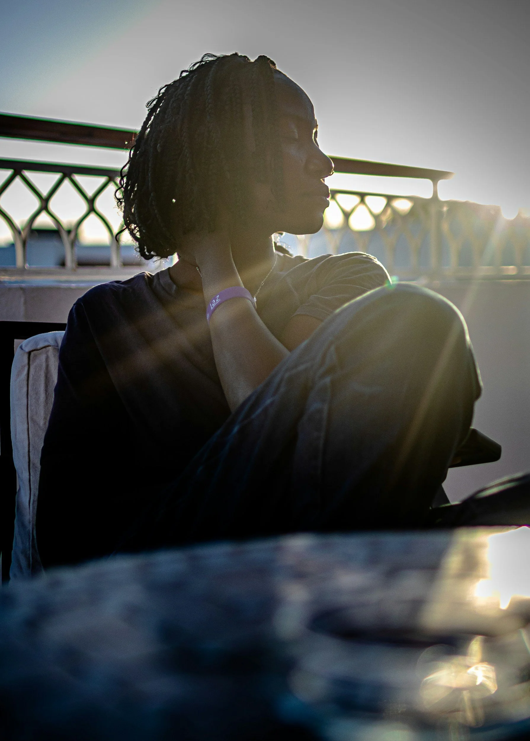 Side profile of a woman with short, curly hair sitting outdoors during sunset, with a metal fence and cityscape in the background.