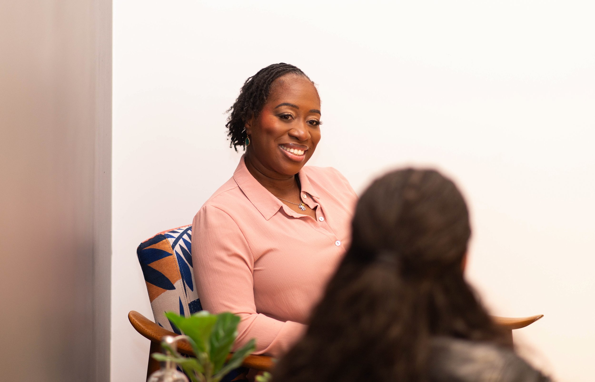 A woman with dark skin, short dreadlocks, and wearing a pink shirt is smiling while talking to a person whose back is visible in the foreground. The setting appears to be an indoor meeting or casual conversation.