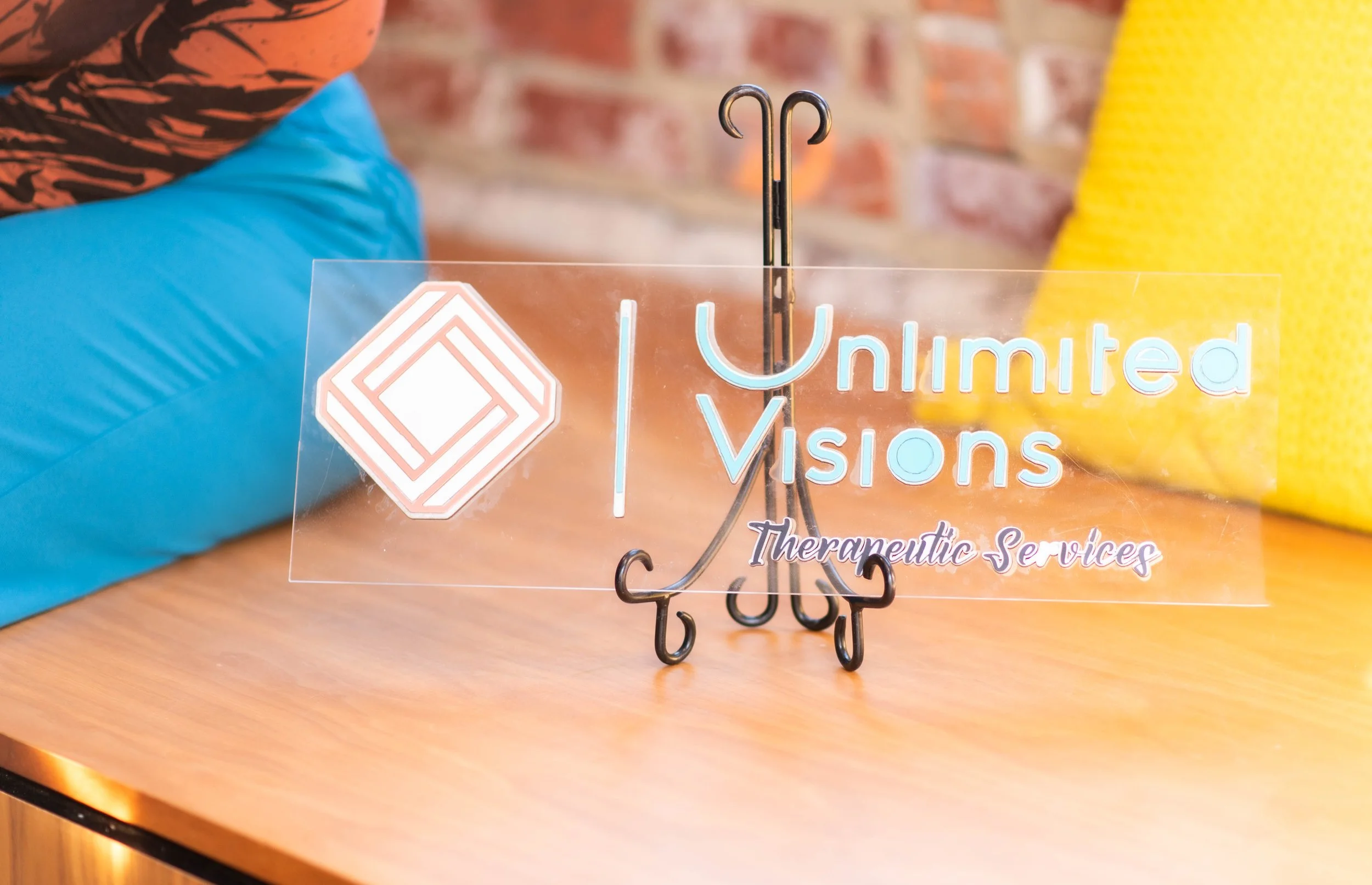 A clear acrylic sign with the words 'Unlimited Visions' and 'Therapeutic Services' printed on it, placed on a wooden table with a decorative black metal stand holding it upright. Behind the sign, there is a brick wall, and part of a person wearing a blue jacket and clothing with orange and black pattern is visible.