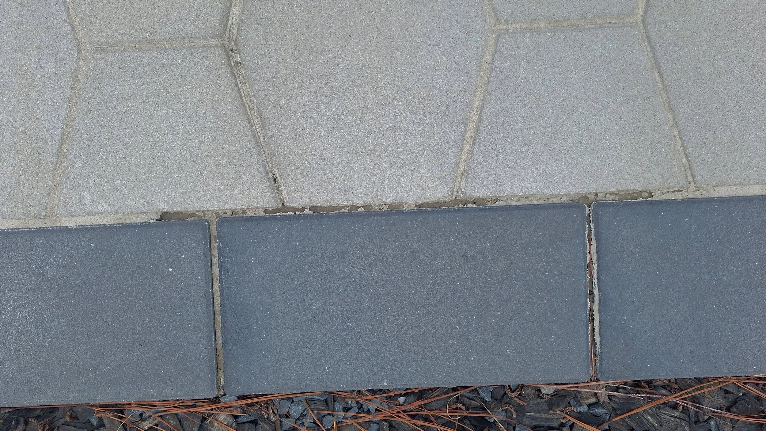 A close-up view of a paved surface showing light gray and dark gray rectangular tiles arranged in a border pattern, with small rocks and dried pine needles along the bottom edge.