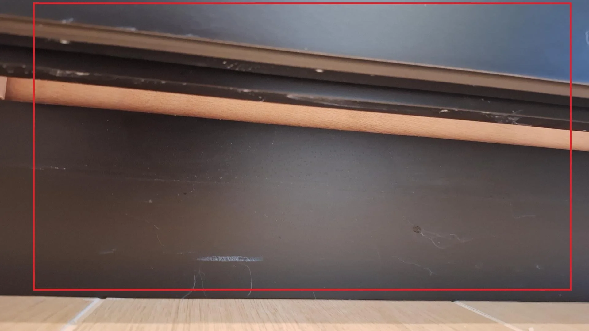 Close-up of a dark-colored surface, possibly a piece of furniture, with some dust and string fibers on it. Above the surface, there are stacked items including a black object, a wooden slat, and a blue item.