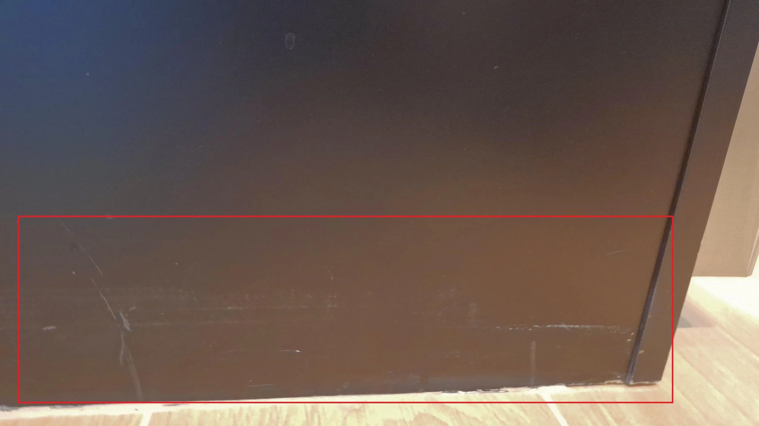 Close-up of a dark-colored wooden or laminate surface, showing some scratches and scuffs, with a portion of a beige or light-colored wall or floor visible at the bottom right corner.