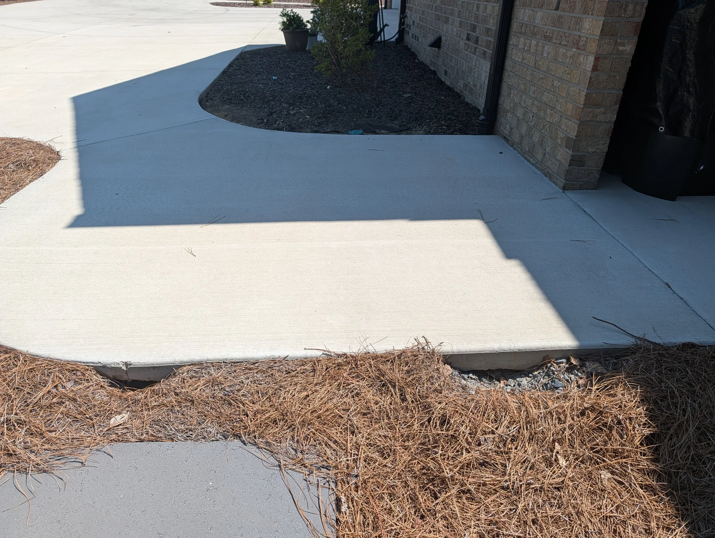 A concrete sidewalk next to a brick house with a small garden bed and a potted plant. Shadows cover part of the sidewalk, and there's a black object leaning against the house.