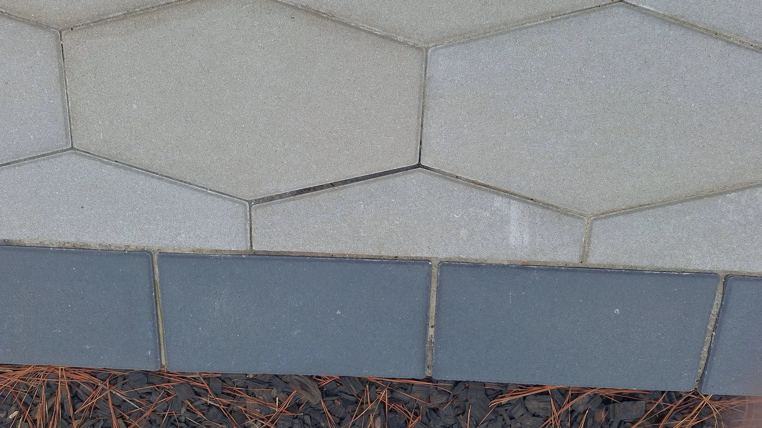 Close-up of a brick wall with alternating light gray and dark gray bricks, surrounded by pine needles and black mulch.