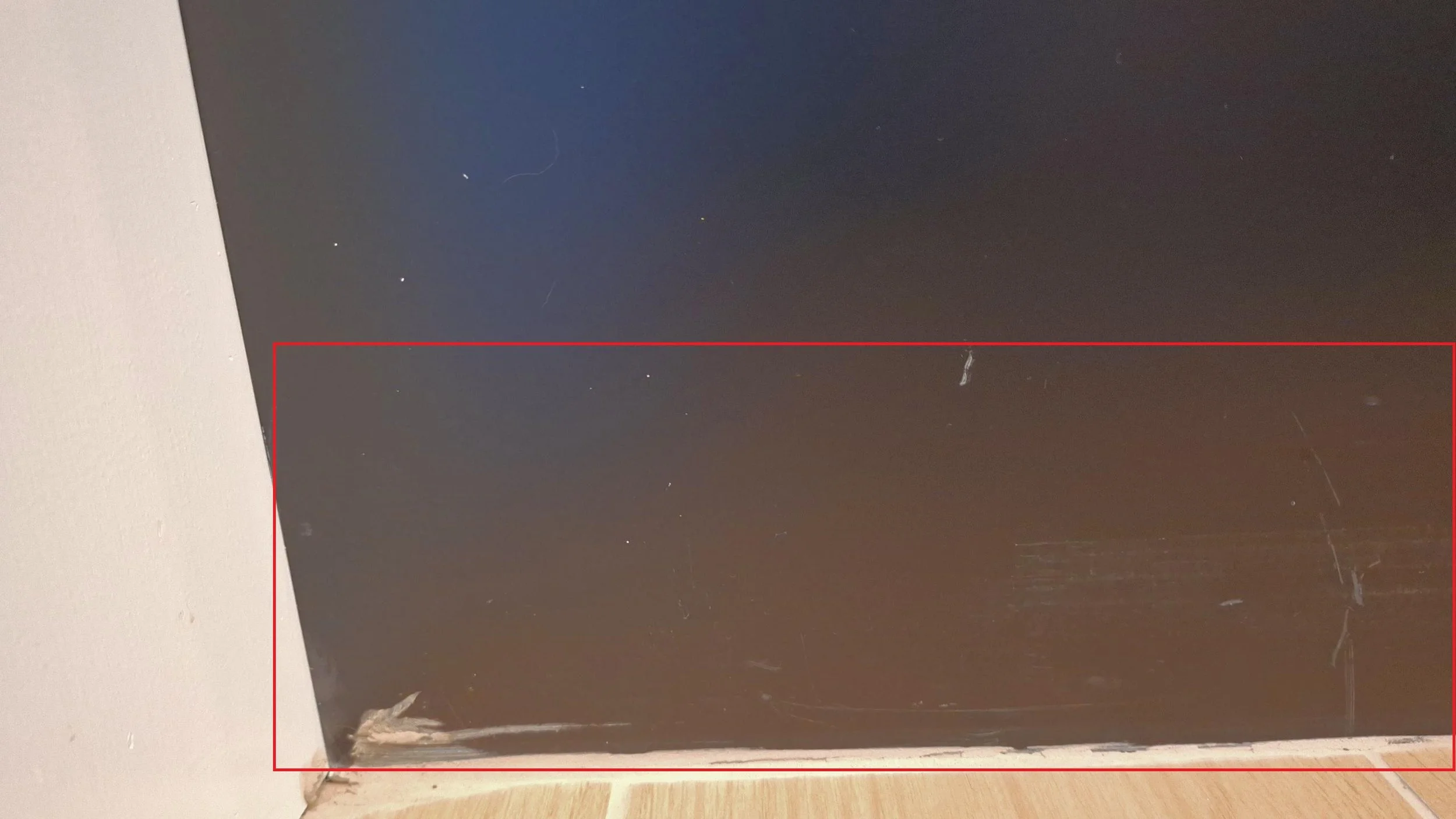 Close-up image of the bottom corner of a black piece of furniture with visible scratches and scuffs, against a white wall and a wooden floor.