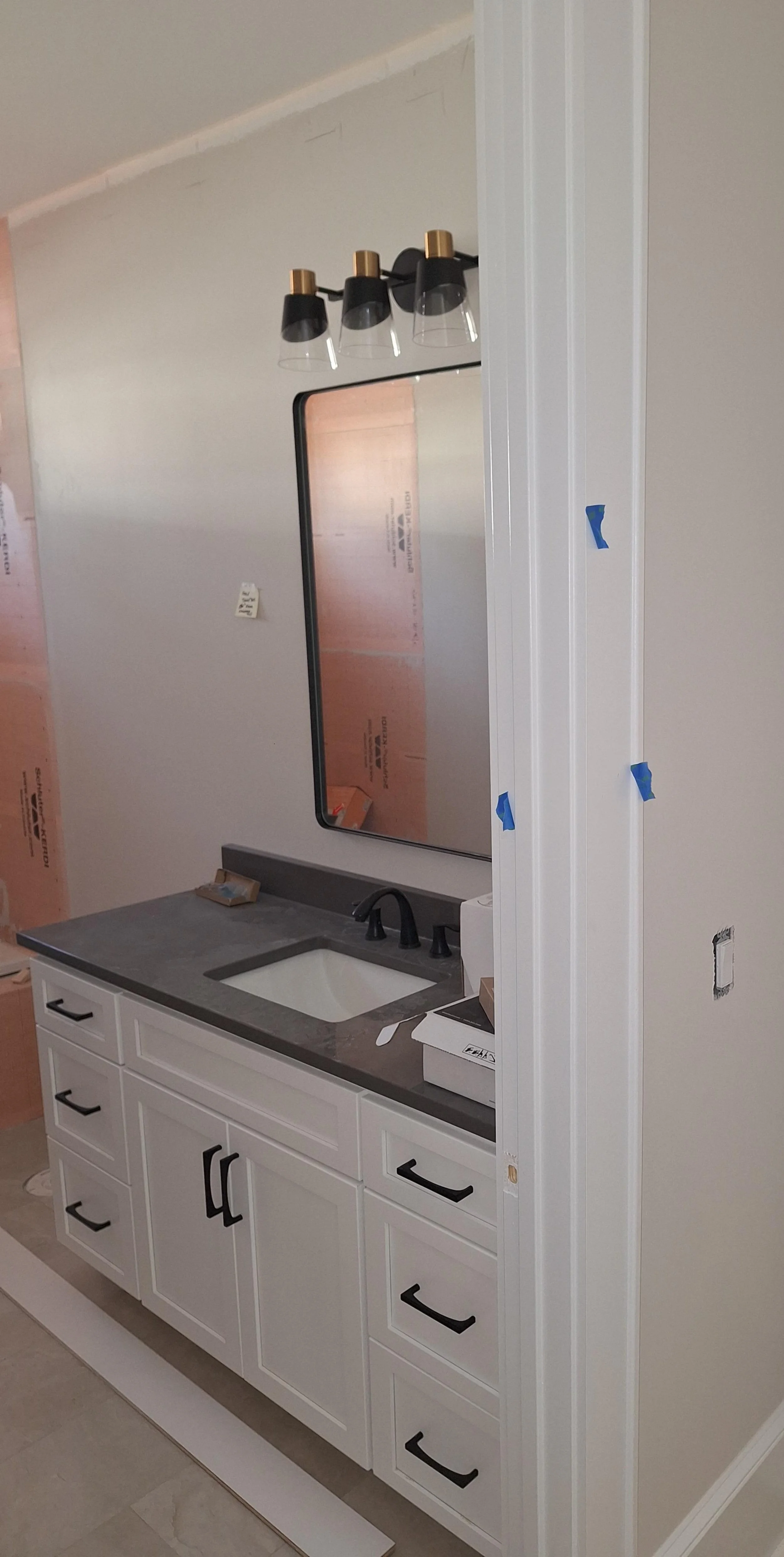 White bathroom vanity with black handles, gray countertop, under-mount sink, black faucet, large mirror, black light fixture with three glass shades, unfinished wall with gaping hole and construction tape.
