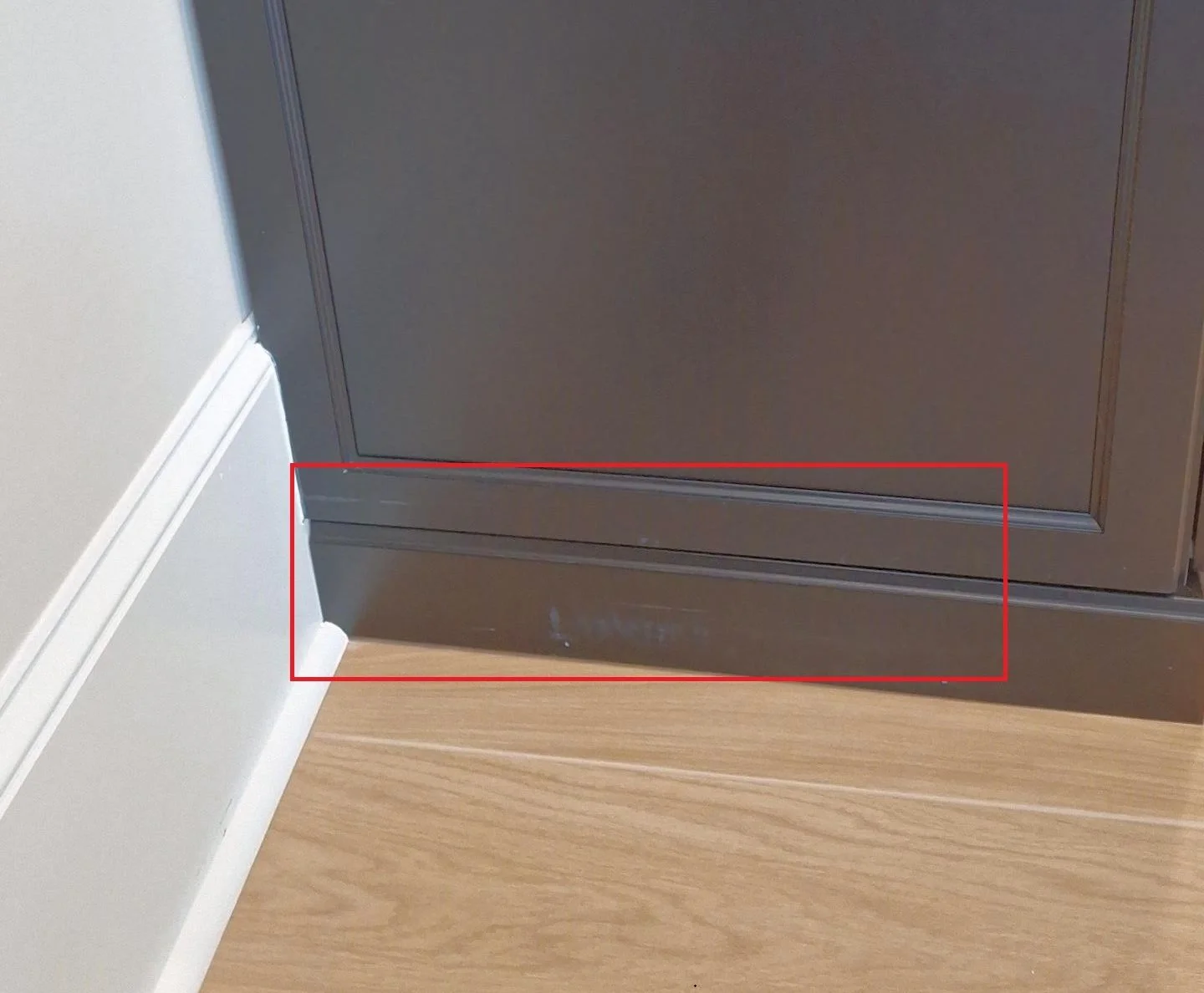 Close-up of the bottom part of a dark-colored cabinet against a beige wall and wooden floor, with a red rectangle highlighting the base of the cabinet and the corner where the wall, baseboard, and floor meet.