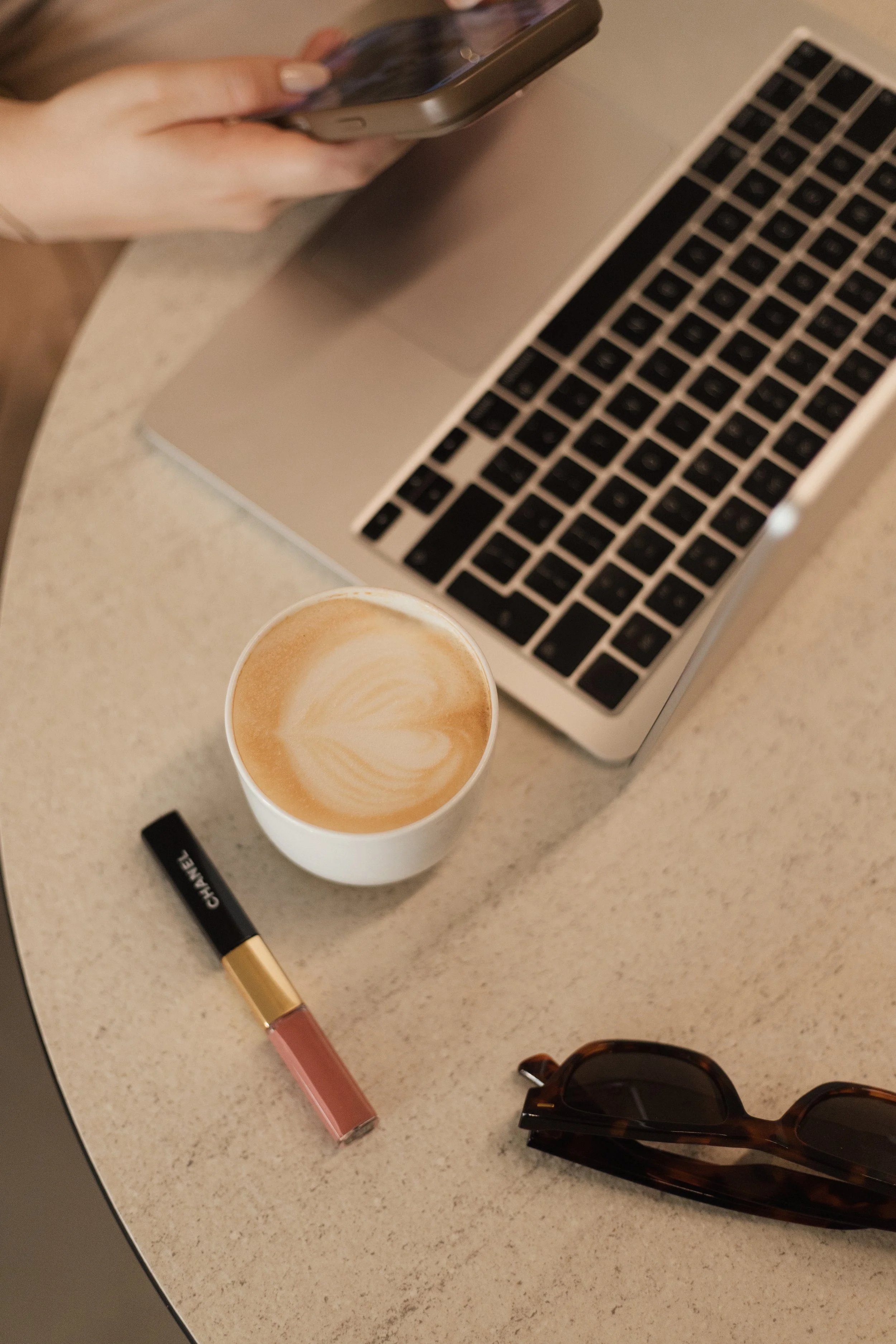 Tasse mit Latte Macchiato, MacBook, Handy, Wasserfalsche, Sonnenbrille, Lippenstift, und andere Accessoires auf einem Tisch.