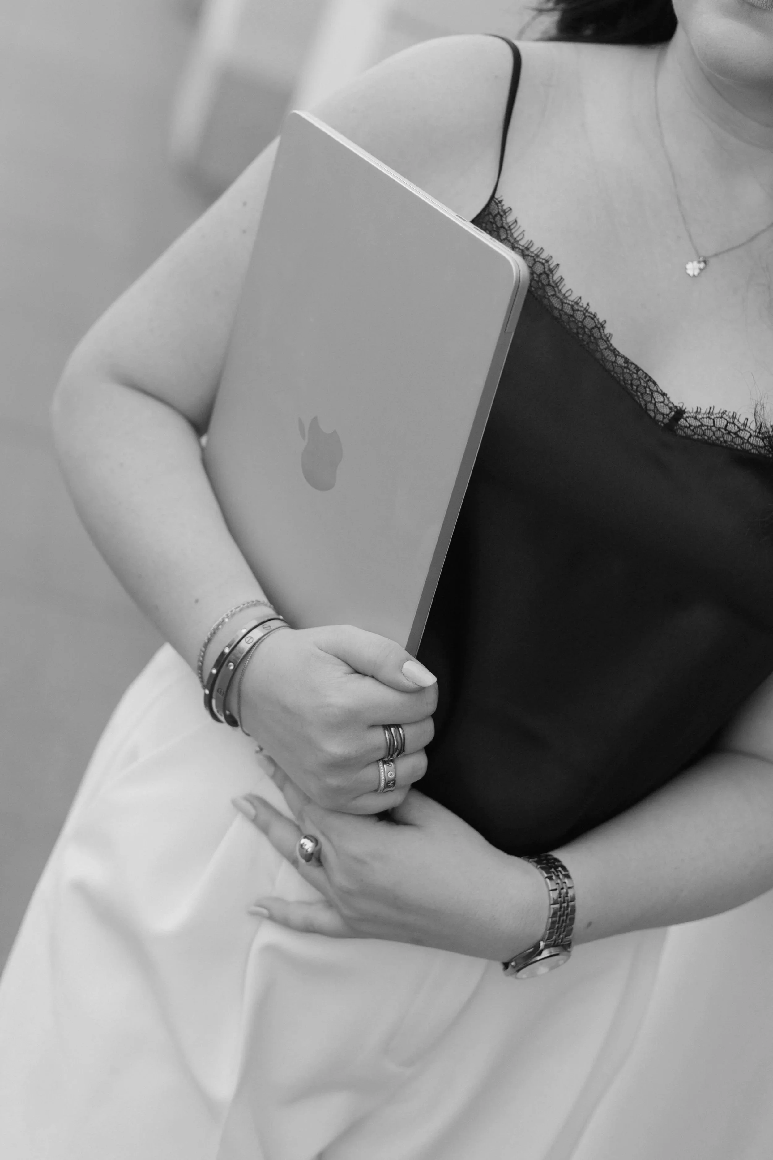 Nahaufnahme eines Frauenarms, die sie trägt mehrere Armbänder und eine Armbanduhr. Sie hält einen Laptop auf ihrer Hüfte. Sie trägt ein schwarzes Oberteil mit Spitze am Rand und eine Halskette mit Anhänger.